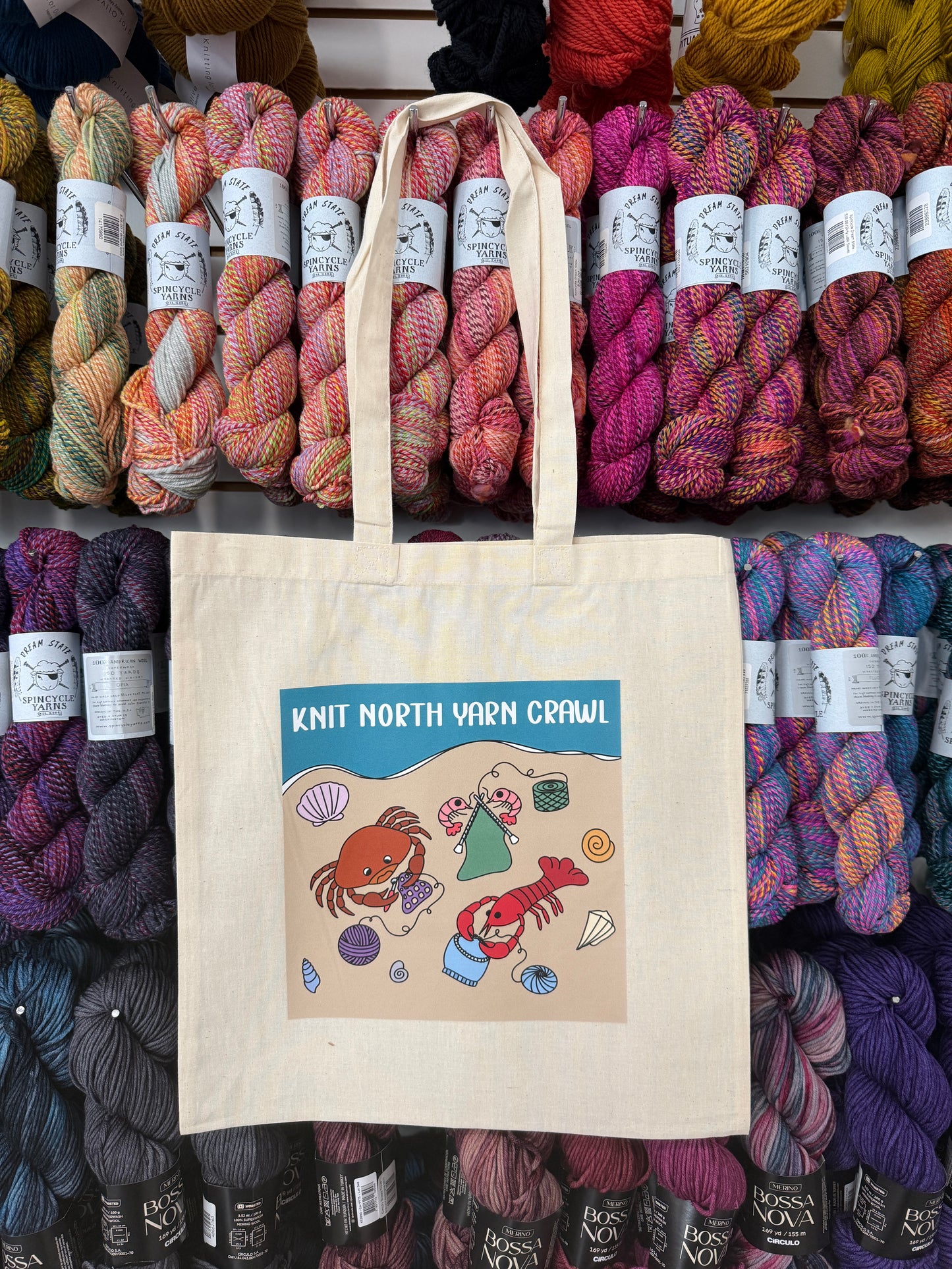 A tote bag featuring a lobster, crab, and shrimp knitting and crocheting design on a beach, with the text 'Knit North Yarn Crawl on a background of colorful yarn skeins.