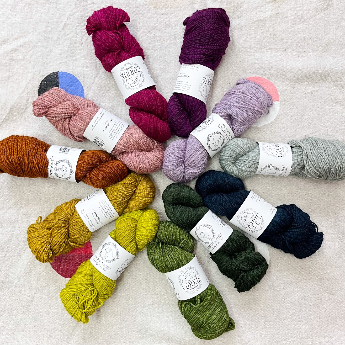 An image of multiple skeins of La Bien Aimee worsted weight yarn in a variety of colors with white labels featuring an image of a sheep and the title ‘Corrie Worsted’ displayed in a pinwheel shape against a canvas background.