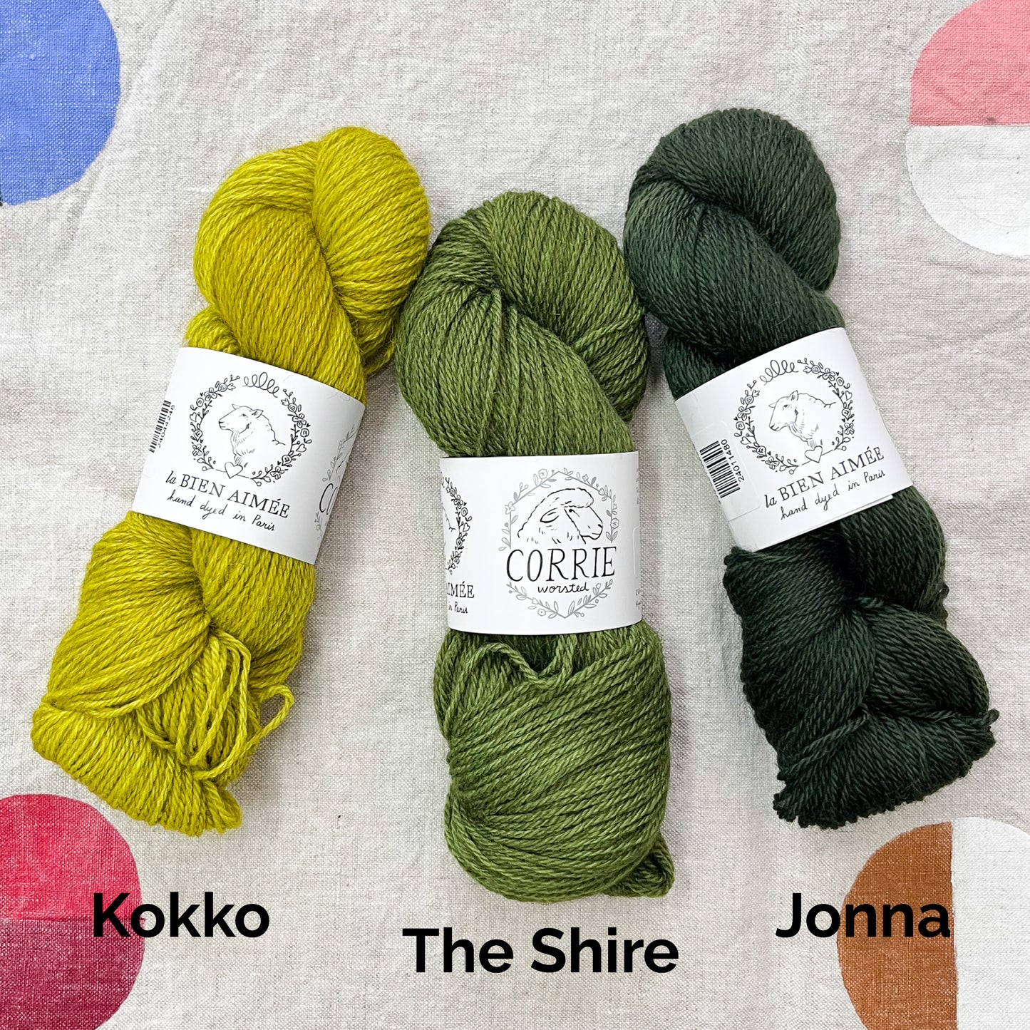 An image of a skein of yellow green yarn on the left, a skein of green yarn in the middle, and a skein of forest green yarn on the right, all with white labels featuring an image of a sheep and the titles ‘La Bien Aimee’ and ‘Corrie Worsted’ against a canvas background.