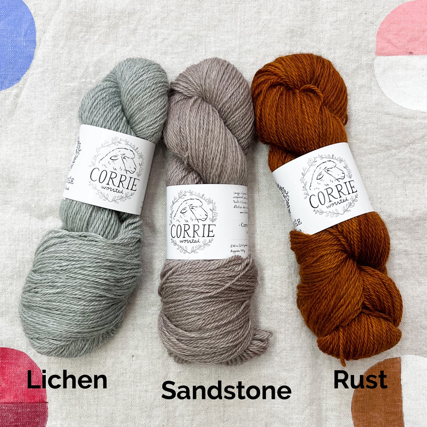 An image of a skein of light gray yarn on the left, a skein of taupe yarn in the middle, and a skein of rust orange yarn on the right, all with white labels featuring an image of a sheep and the title ‘Corrie Worsted’ against a canvas background.