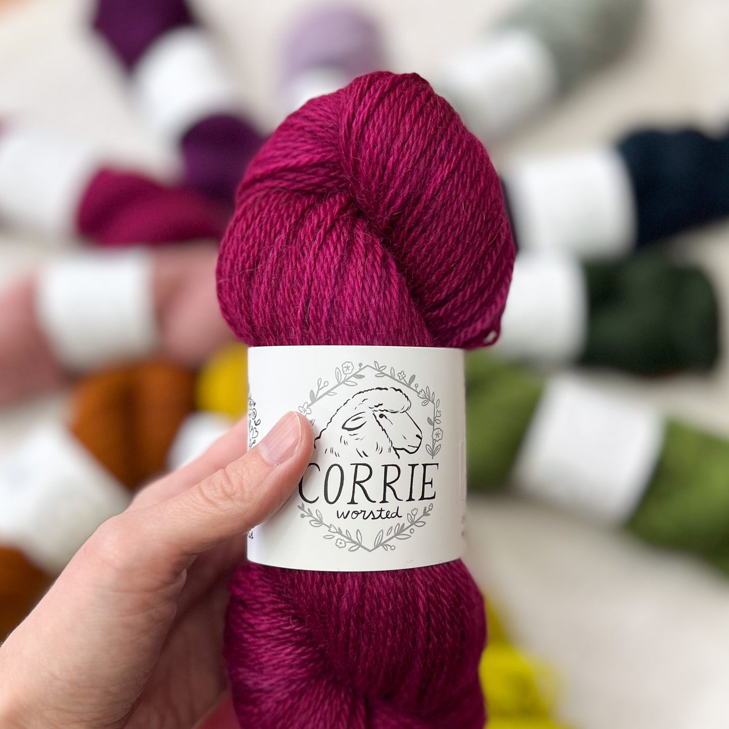 An image of a skein of La Bien Aimee worsted weight yarn in a vibrant fuchsia color, with a white label featuring a sheep and the title ‘Corrie Worsted’ held in a hand against a blurred yarn display background.