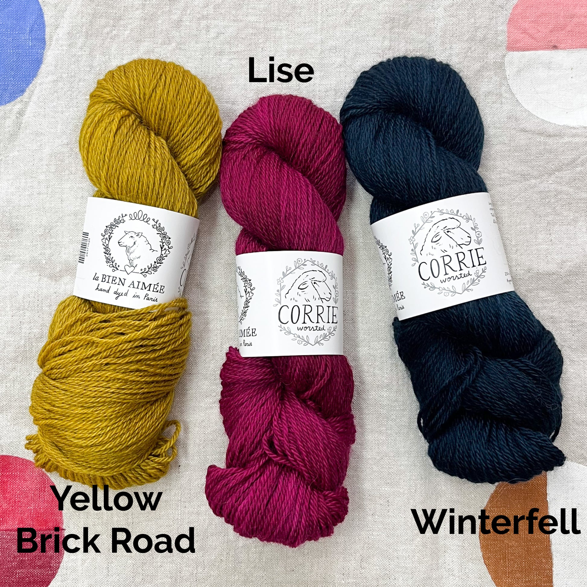 An image of a skein of mustard yellow yarn on the left, a skein of fuchsia yarn in the middle, and a skein of navy blue yarn on the right, all with white labels featuring an image of a sheep and the titles ‘La Bien Aimee’ and ‘Corrie Worsted’ against a canvas background.