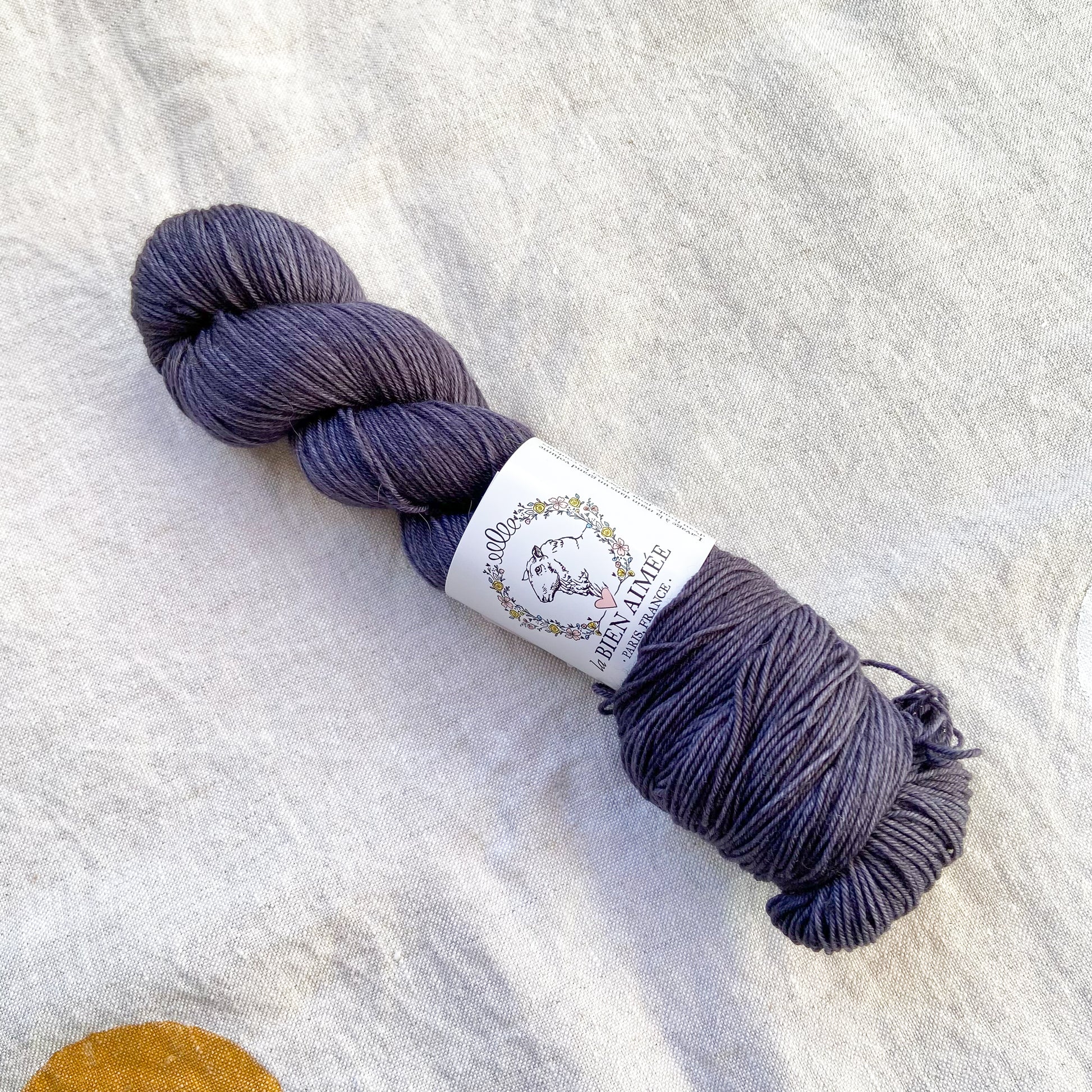 An image of a skein of sock yarn in a charcoal gray color with a white ‘La Bien Aimee’ label against a canvas background.