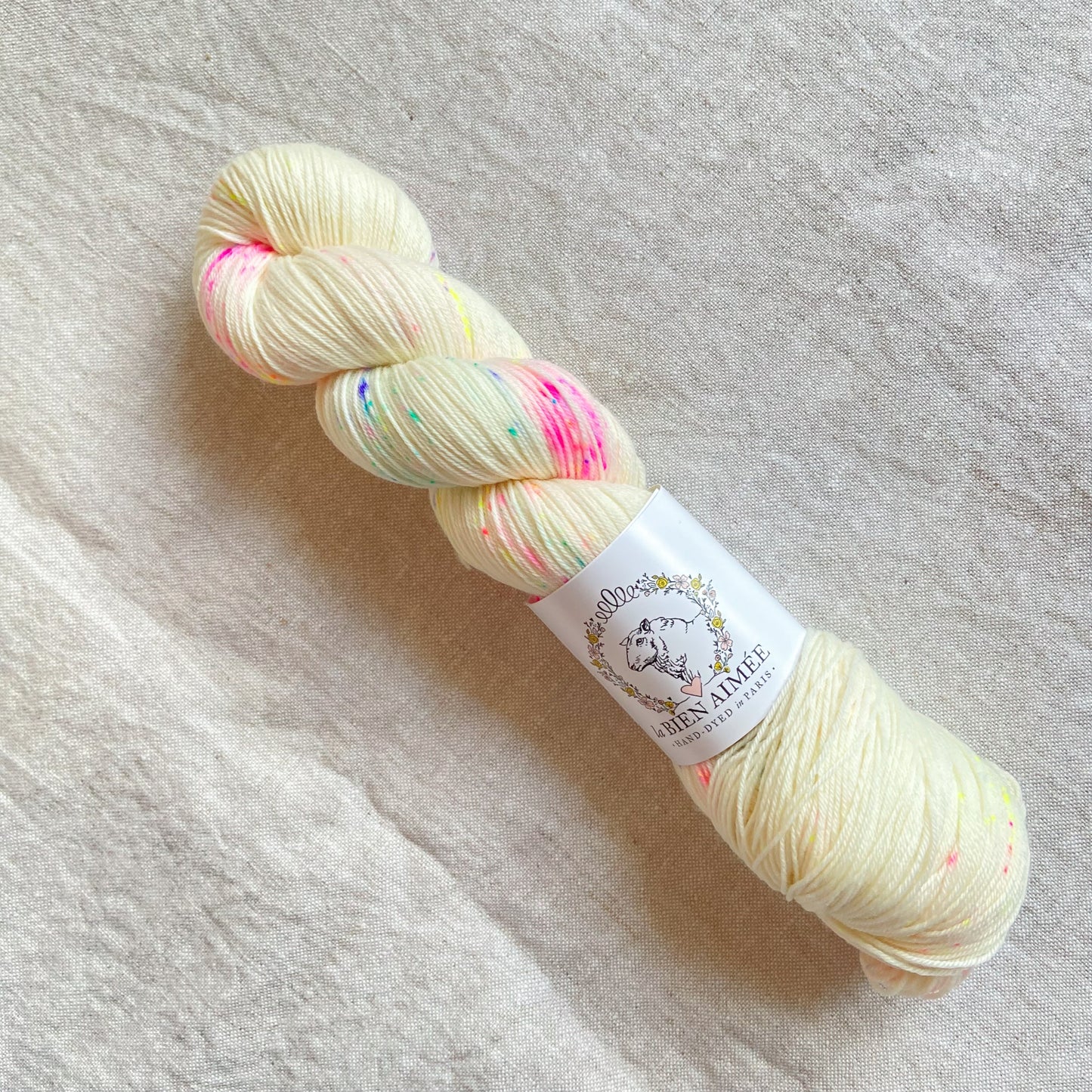 An image of a skein of sock yarn in a cream color speckled with rainbow colors, with a white ‘La Bien Aimee’ label against a canvas background.