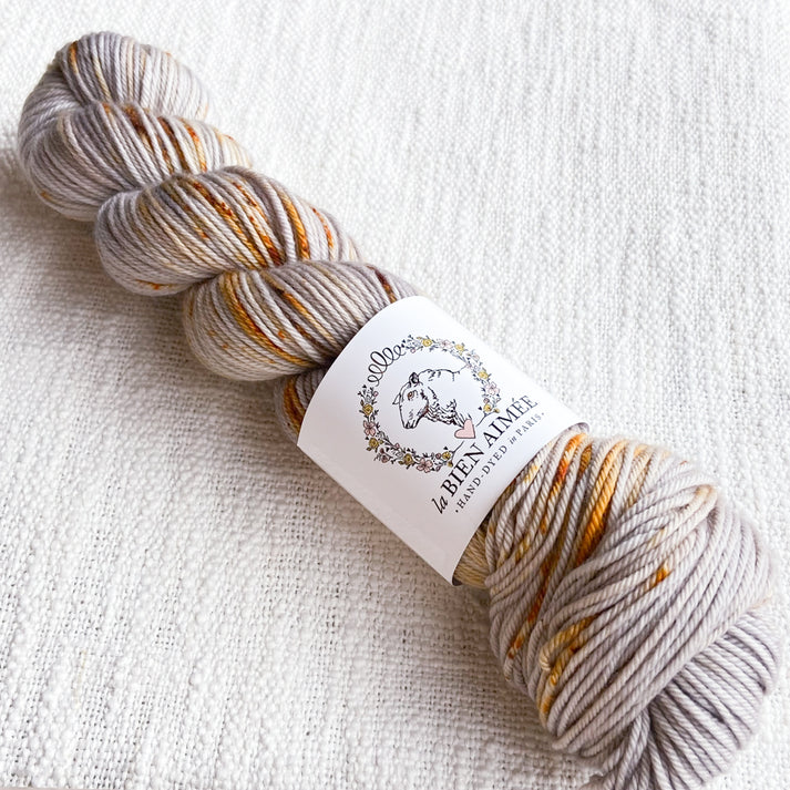 An image of a skein of sock yarn in a taupe color with speckles of orange and yellow, with a white ‘La Bien Aimee’ label against a canvas background.