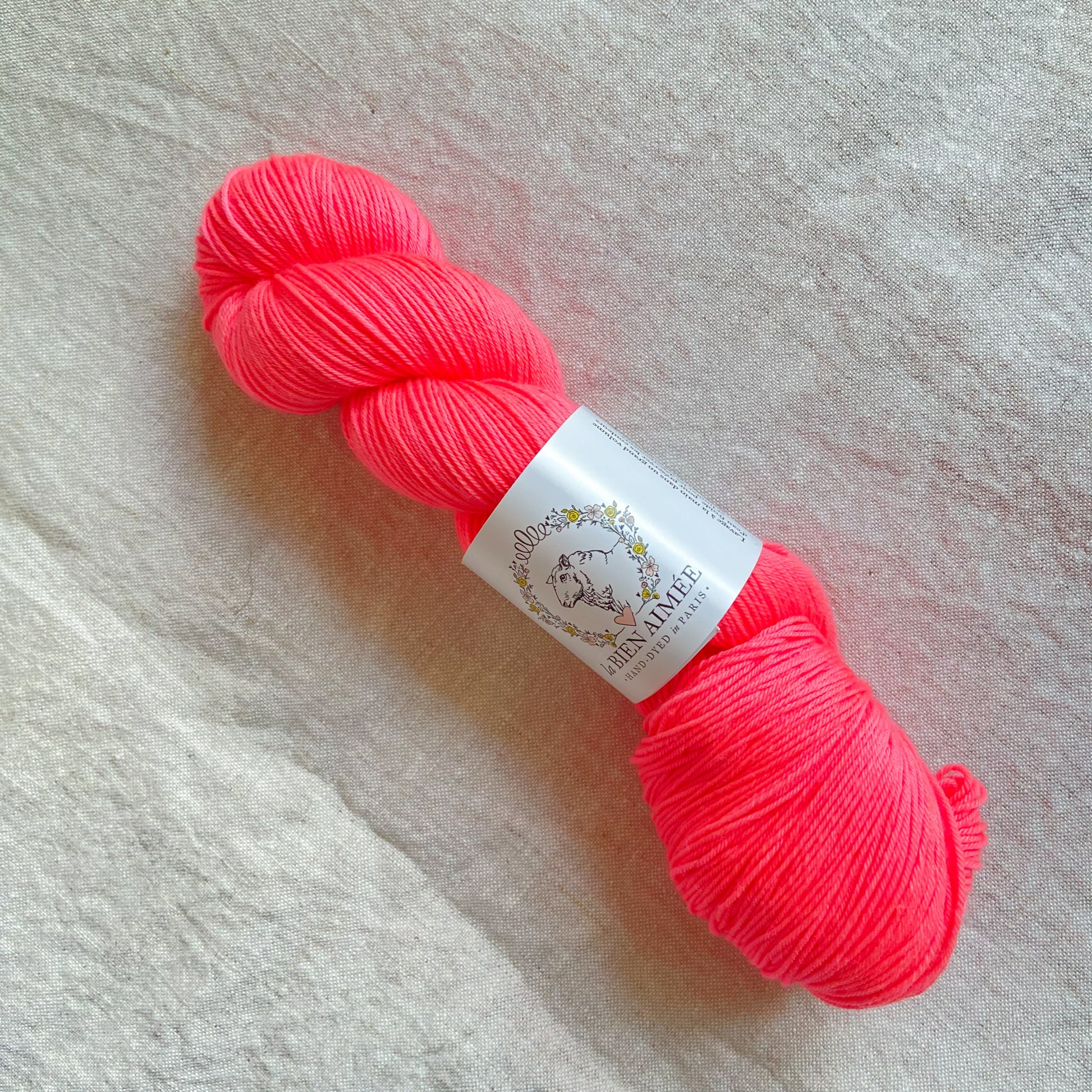 An image of a skein of sock yarn in a neon pink color, with a white ‘La Bien Aimee’ label against a canvas background.