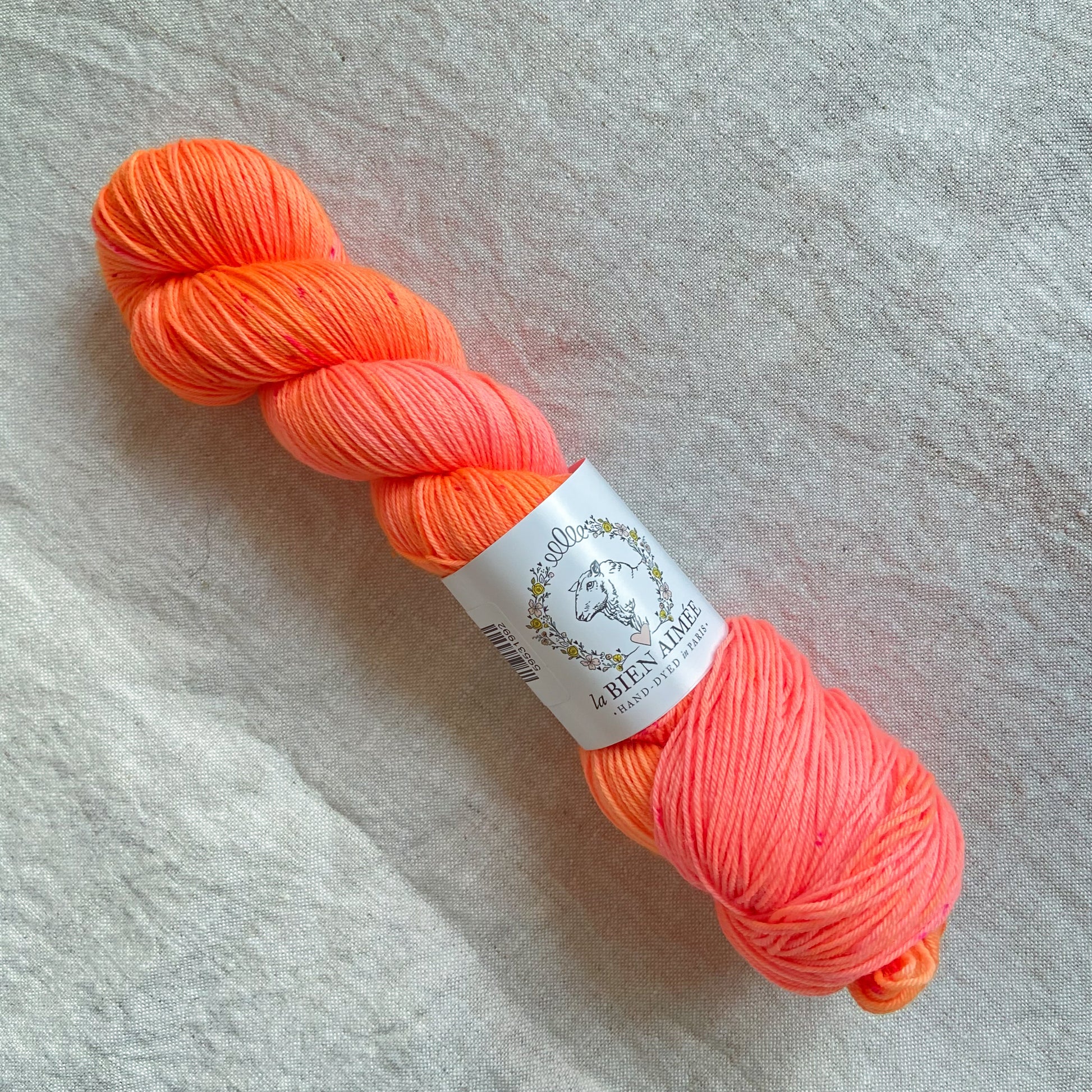 An image of a skein of sock yarn in neon shades of orange and pink, with a white ‘La Bien Aimee’ label against a canvas background.
