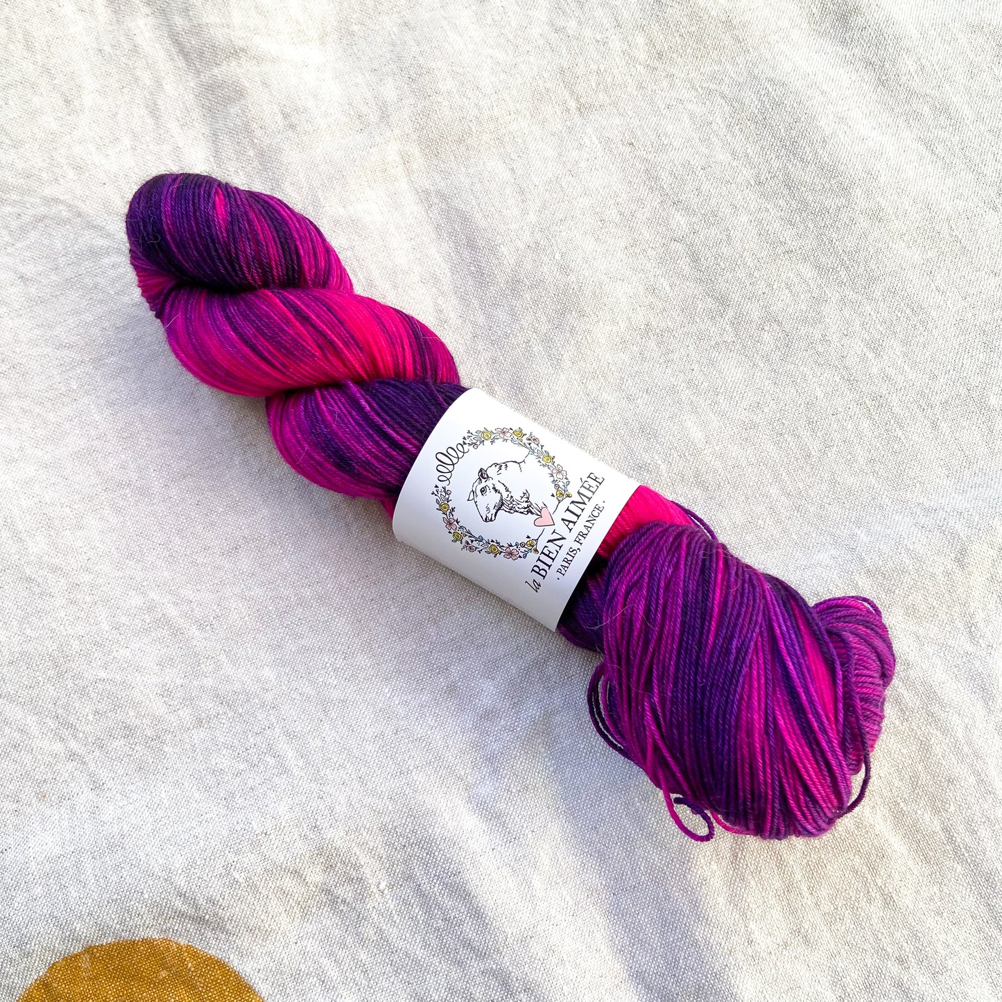 An image of a skein of sock yarn in vibrant shades of pink and purple with a white ‘La Bien Aimee’ label against a canvas background.