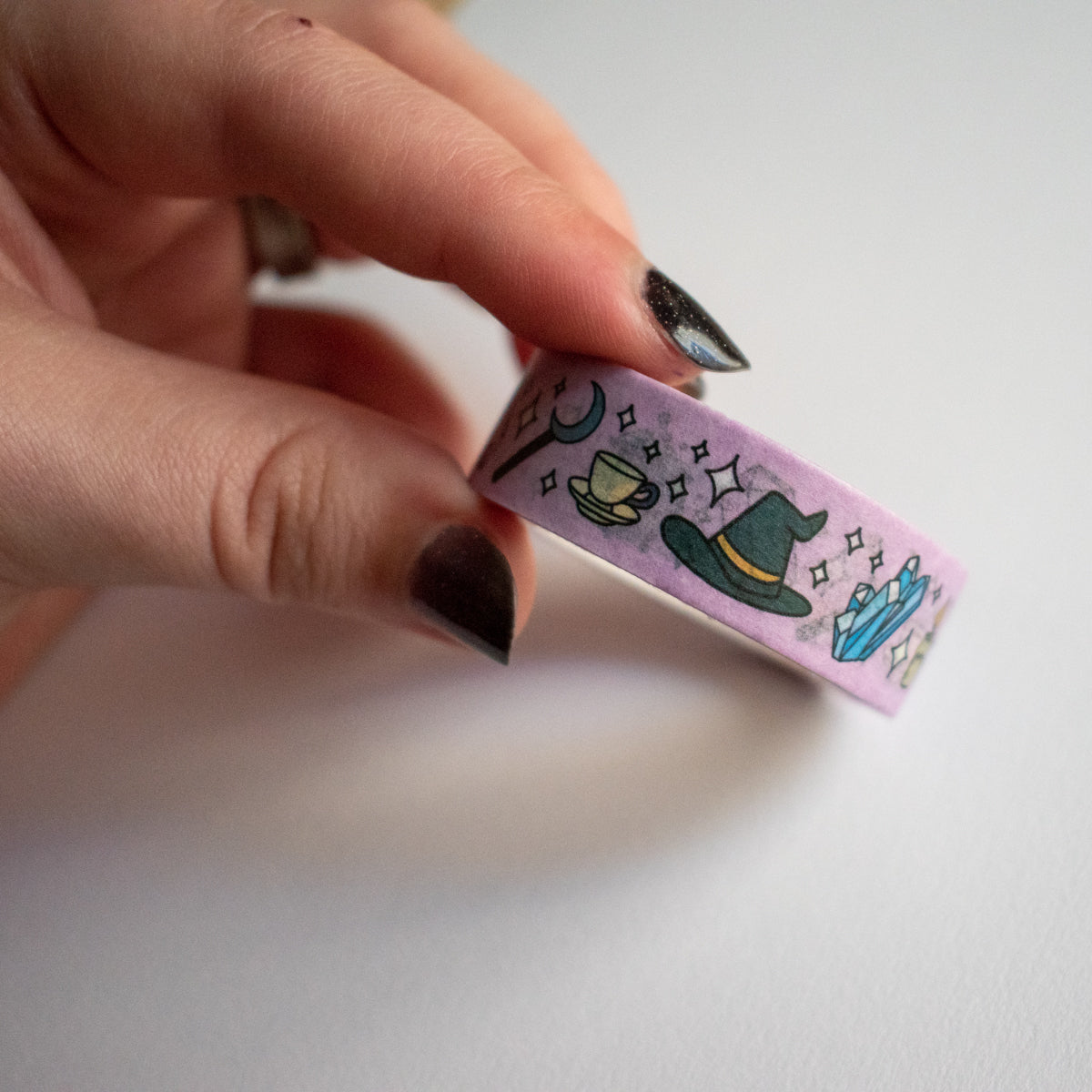 A hand with black painted nails holding a pink roll of washi tape with witchy items on it.