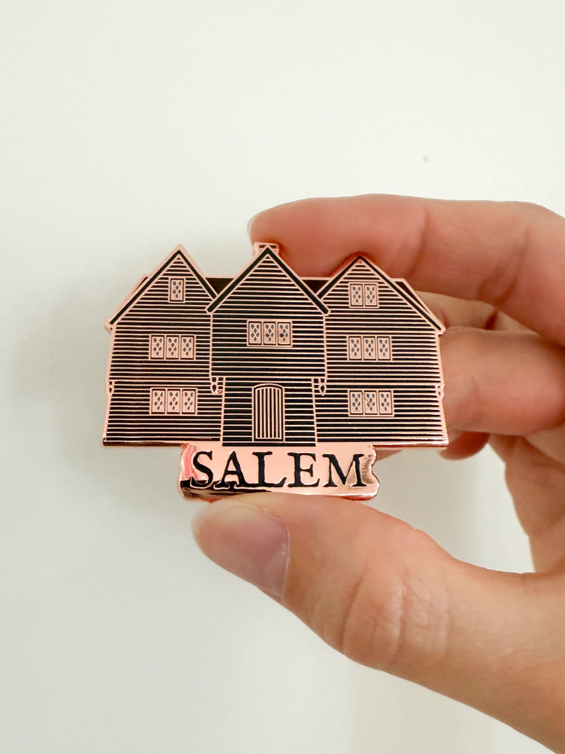 Hand holding a rose gold enamel pin of an illustration of the Witch House in Salem Massachusetts