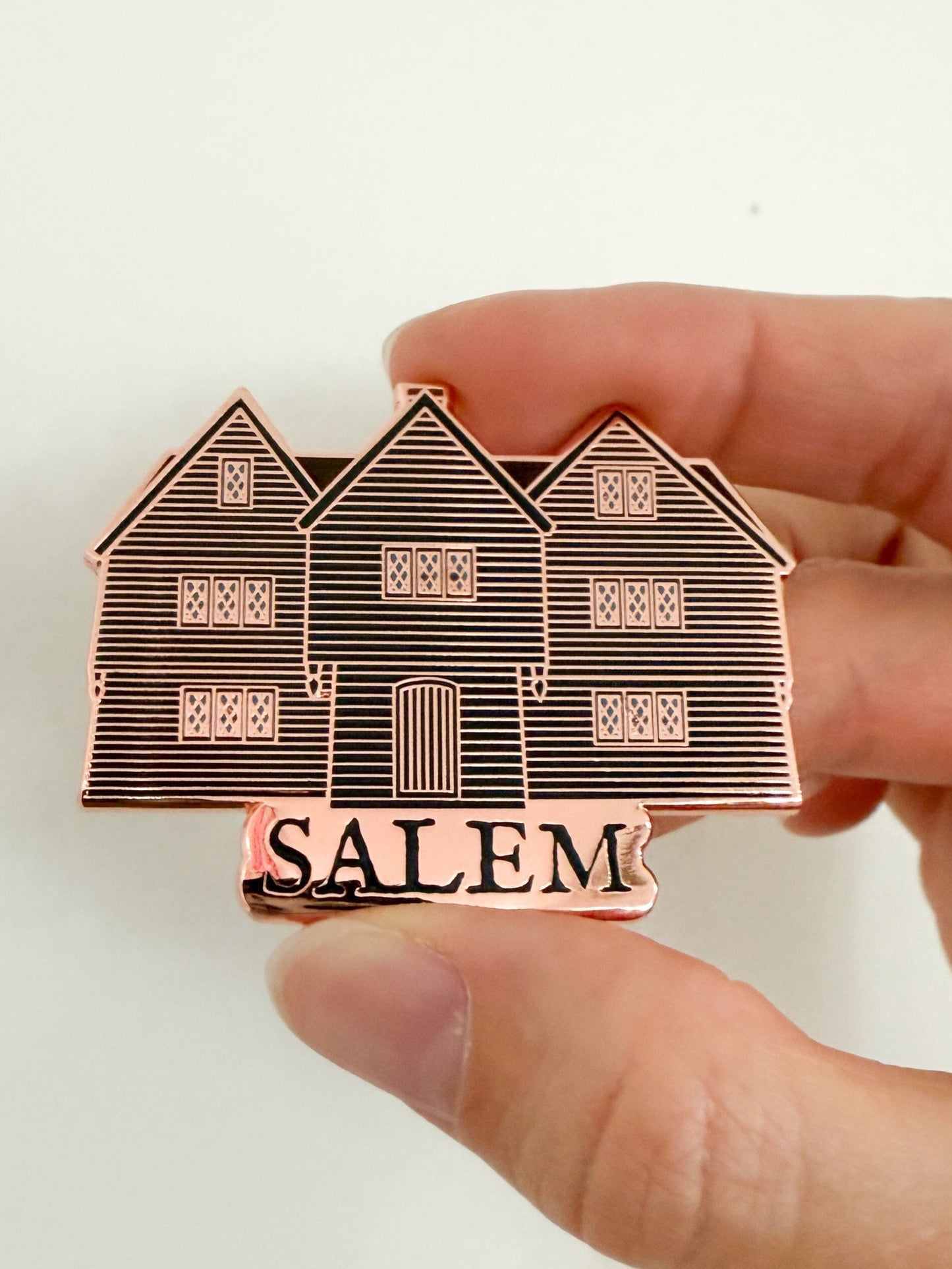 Hand holding a rose gold enamel pin of an illustration of the Witch House in Salem Massachusetts