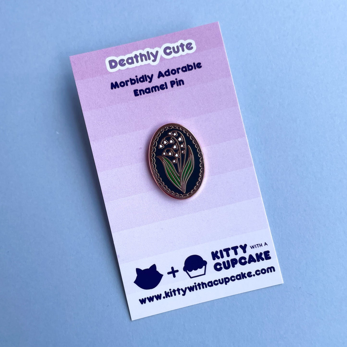 An oval shaped enamel pin with a lily of the valley flower on it on a pink card.