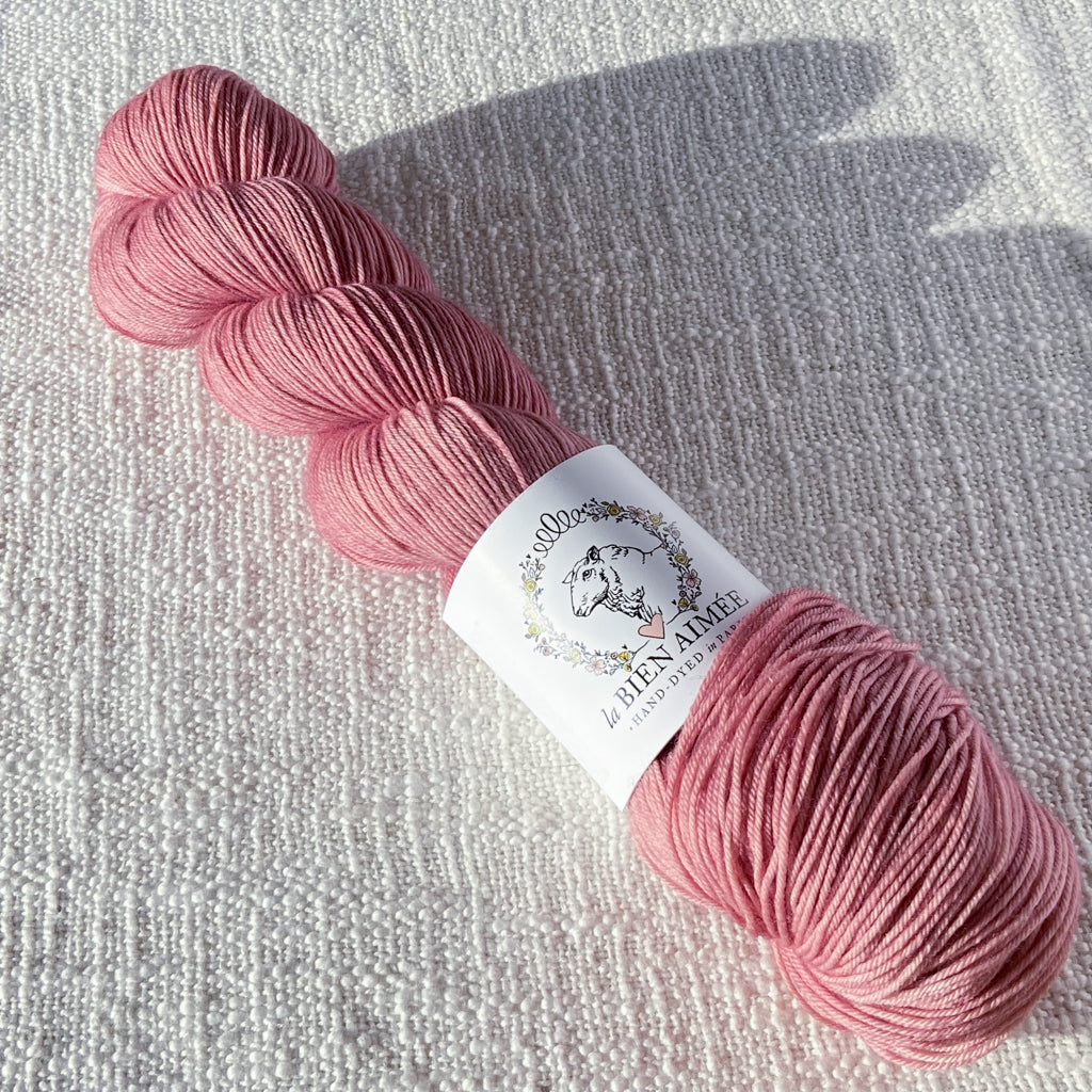 An image of a skein of sock yarn in a rose pink color with a white ‘La Bien Aimee’ label against a canvas background.