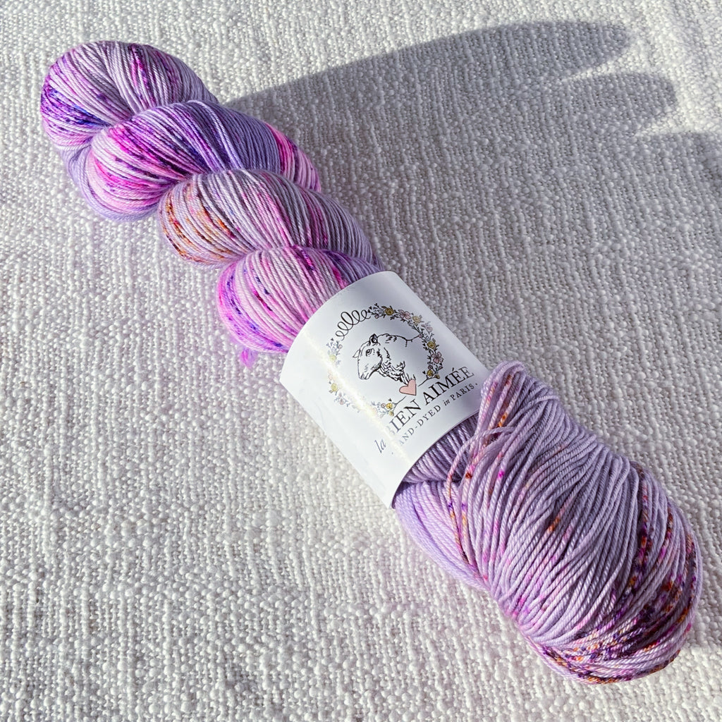 An image of a skein of sock yarn in a light purple base with speckles of pink and orange, with a white ‘La Bien Aimee’ label against a canvas background.