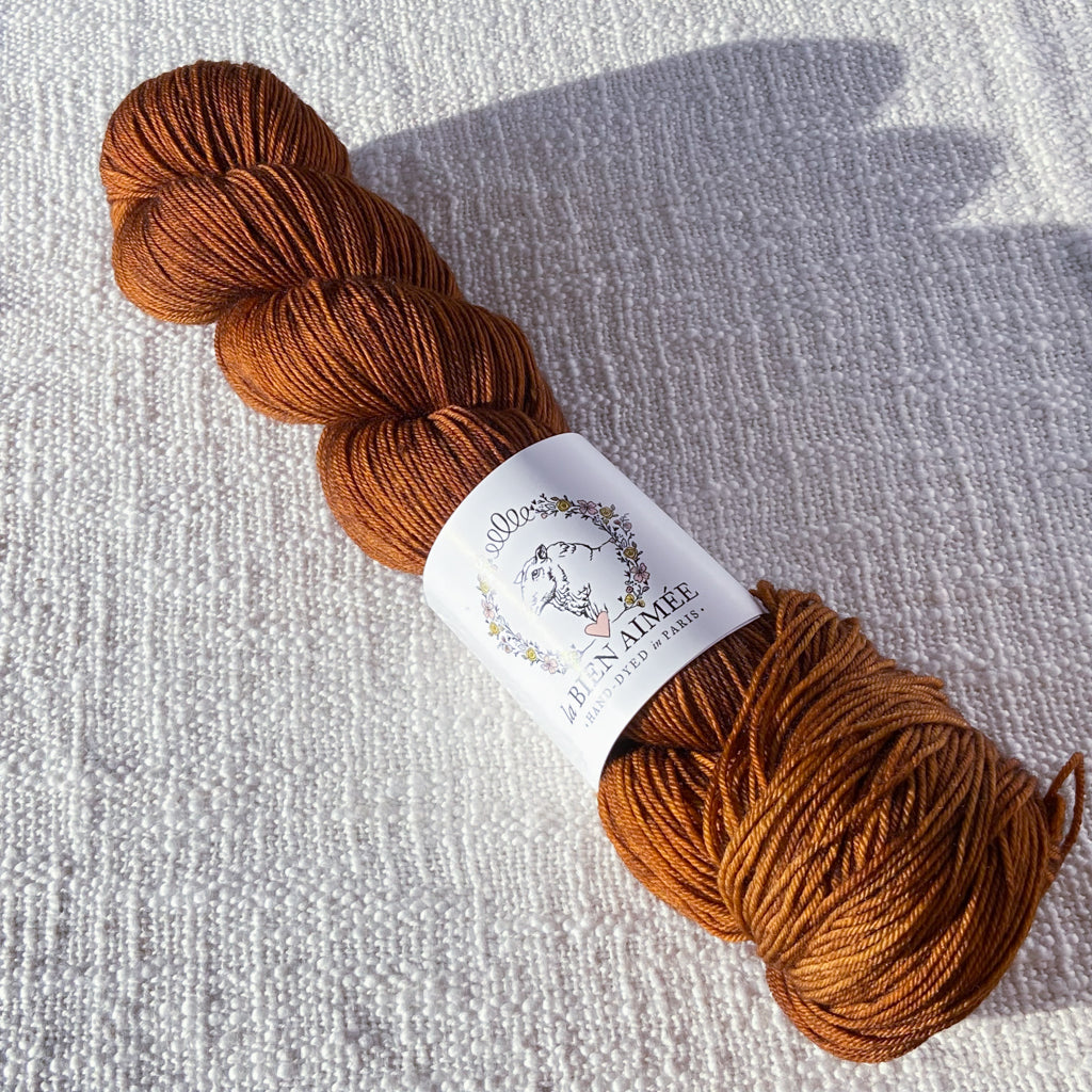 An image of a skein of sock yarn in a light brown color, with a white ‘La Bien Aimee’ label against a canvas background.