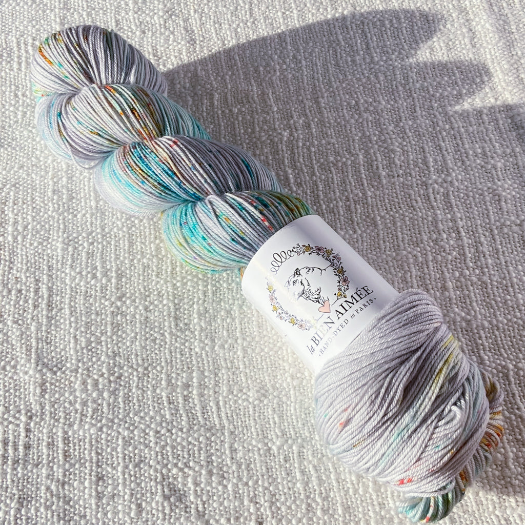 An image of a skein of sock yarn in a light gray base with speckles of blue and orange, with a white ‘La Bien Aimee’ label against a canvas background.