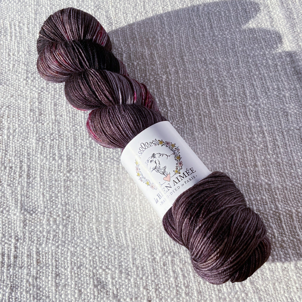 An image of a skein of sock yarn in shades of charcoal, mauve, and pink with a white ‘La Bien Aimee’ label against a canvas background.