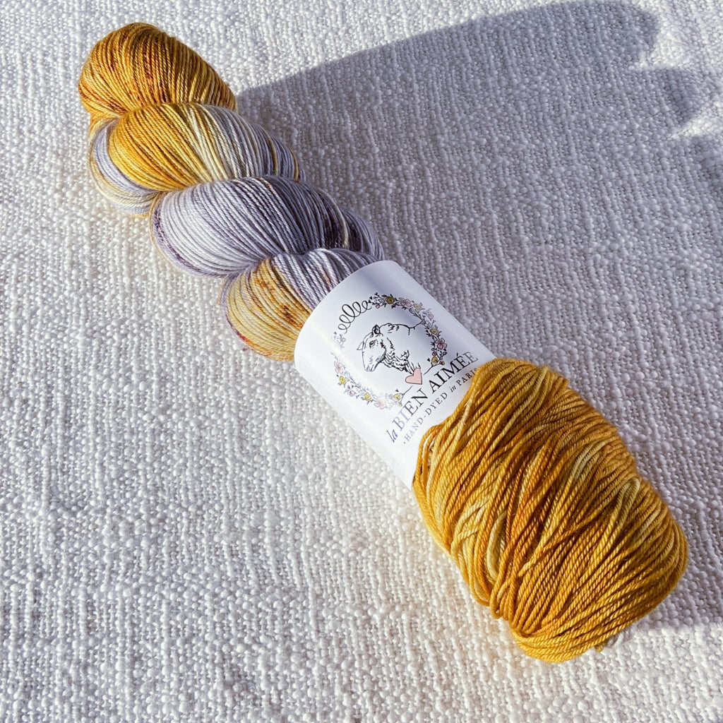 An image of a skein of sock yarn in shades of yellow and gray, with a white ‘La Bien Aimee’ label against a canvas background.