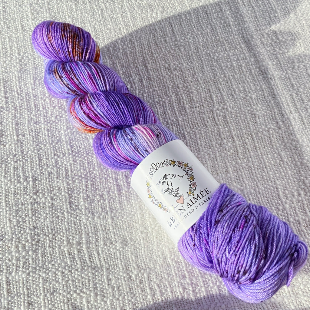 An image of a skein of sock yarn in a bright purple color with speckles of pink and orange, with a white ‘La Bien Aimee’ label against a canvas background.