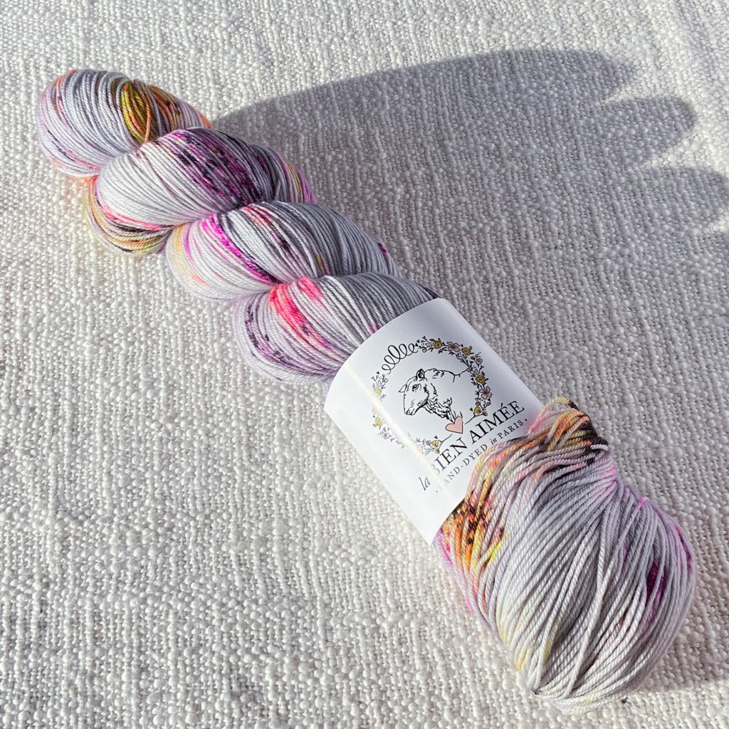 An image of a skein of sock yarn in a light gray color with speckles of pink, purple, and yellow, with a white ‘La Bien Aimee’ label against a canvas background.