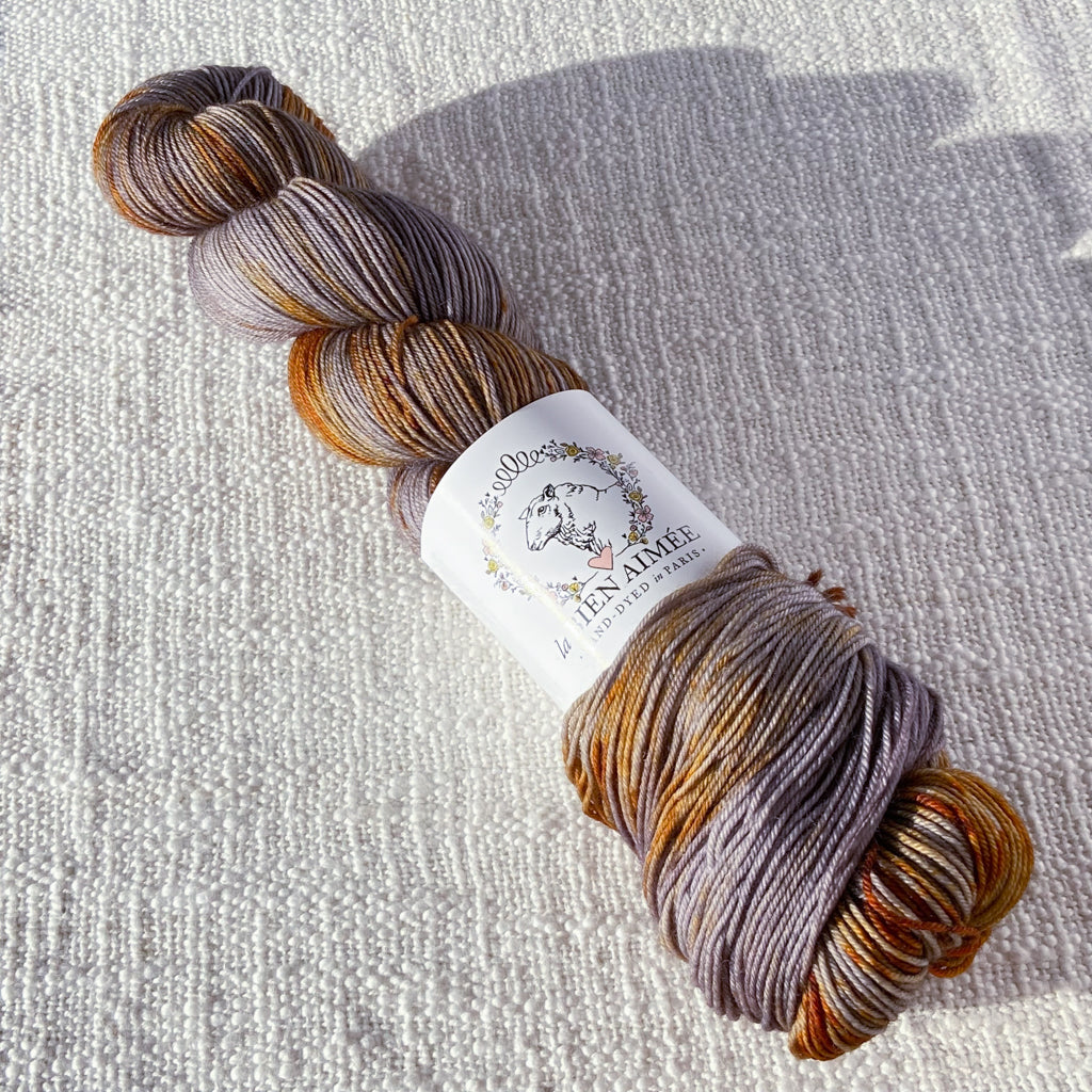 An image of a skein of sock yarn in shades of mauve and rust orange, with a white ‘La Bien Aimee’ label against a canvas background.