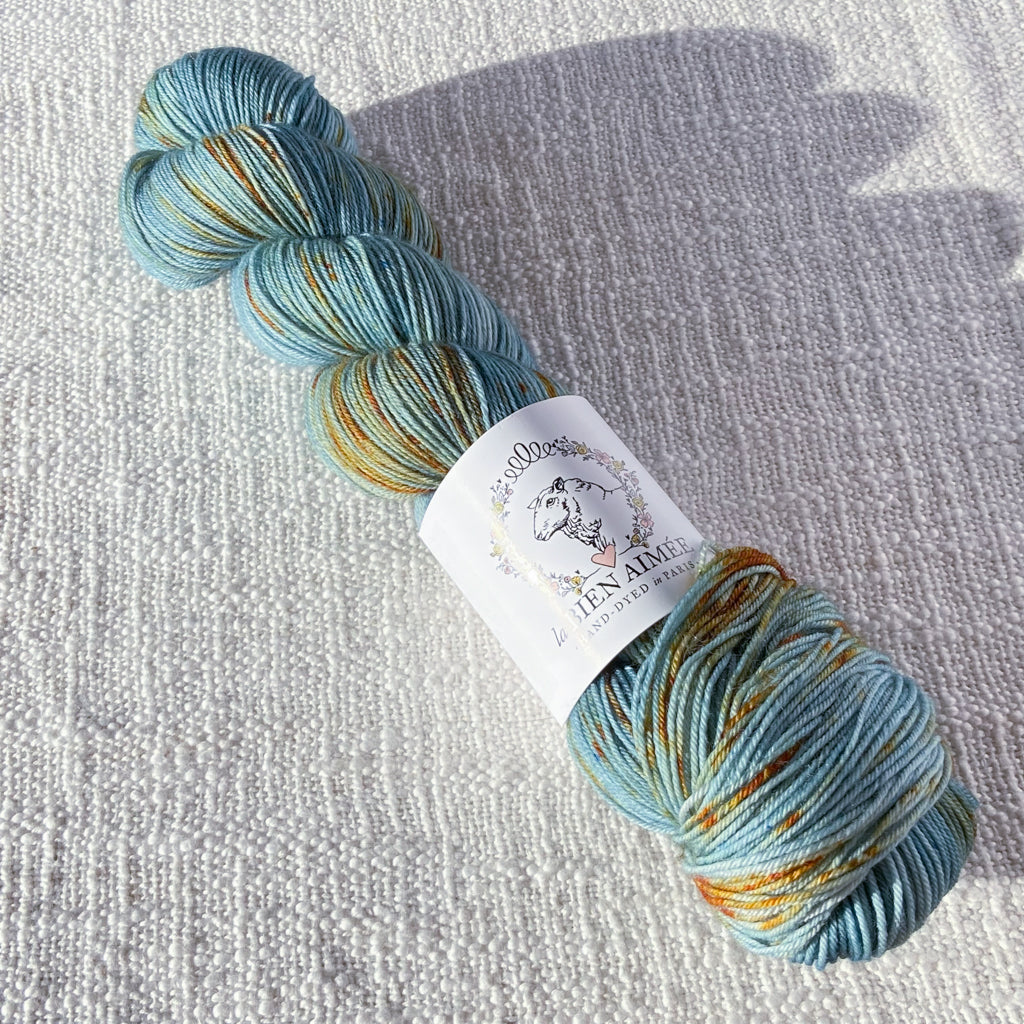An image of a skein of sock yarn in a light blue color with speckles of orange, with a white ‘La Bien Aimee’ label against a canvas background.