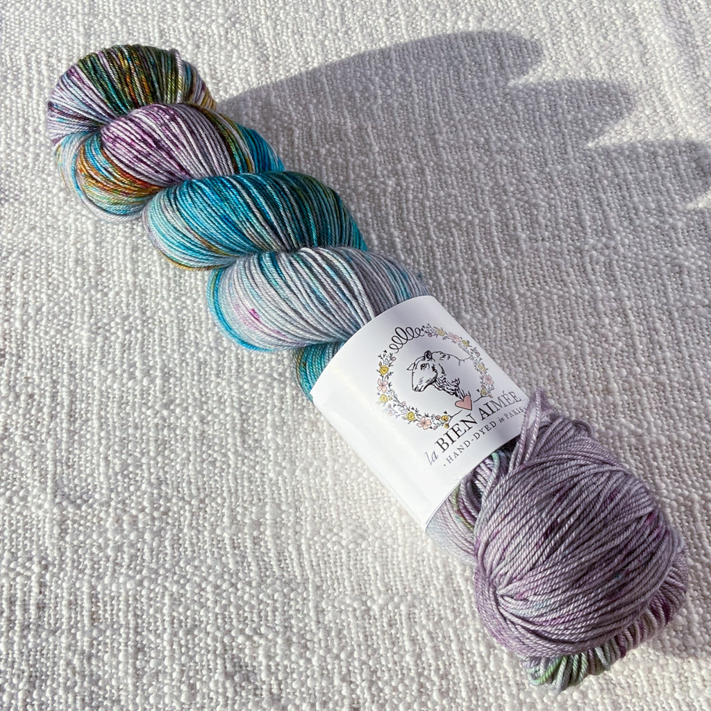 An image of a skein of sock yarn in a shades of mauve, blue, green, and orange, with a white ‘La Bien Aimee’ label against a canvas background.