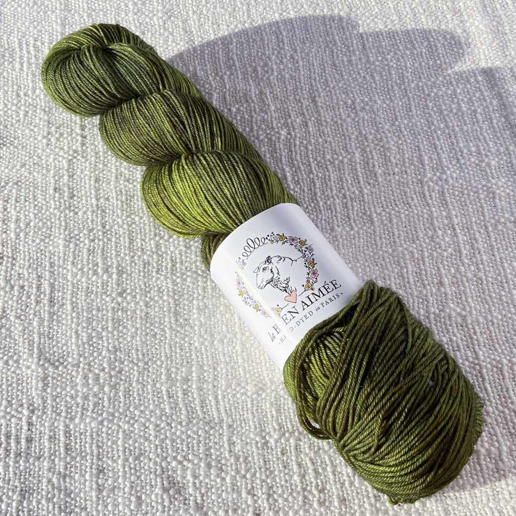 An image of a skein of sock yarn in a forest green color. with a white ‘La Bien Aimee’ label against a canvas background.