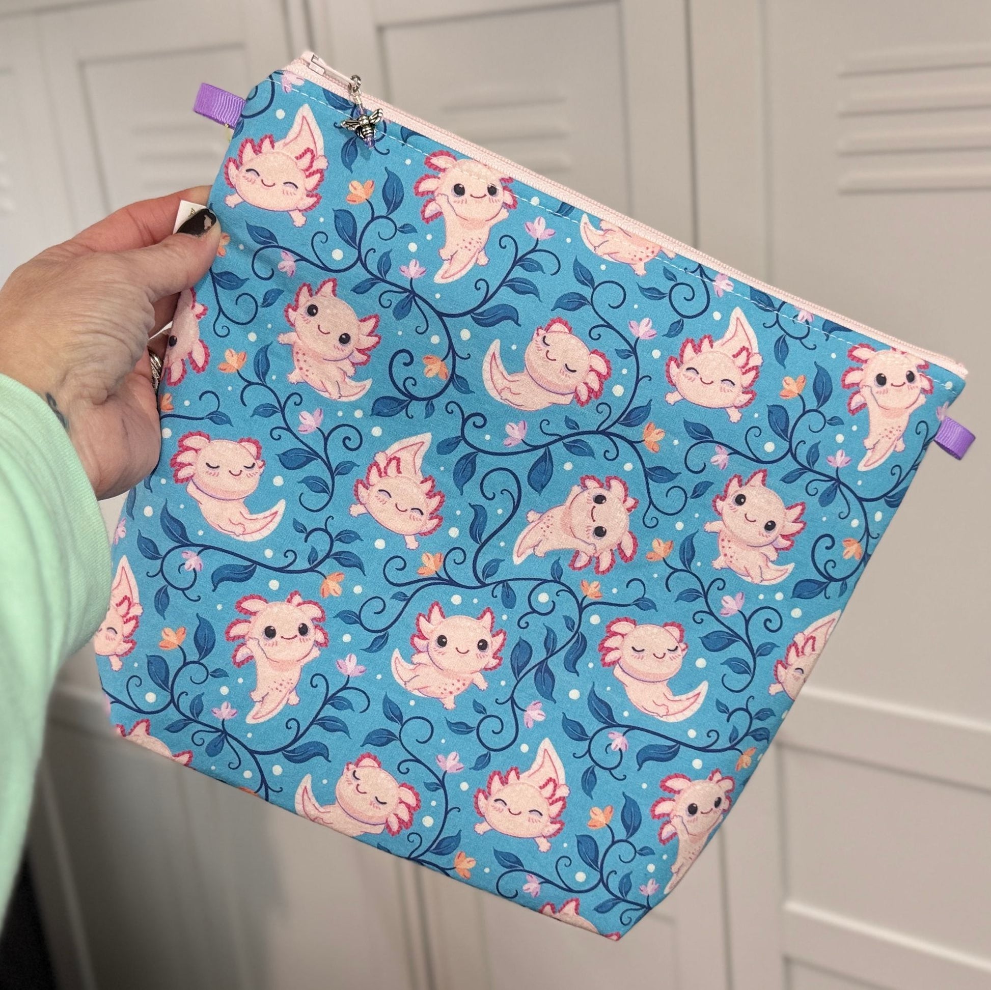 Zippered knitting project with a cute axolotl pattern ob a blue background held by a hand against a white background