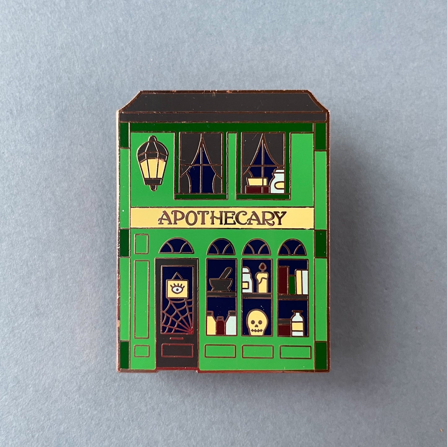 An enamel pin shaped like an Apothecary store front on top of a grey background.
