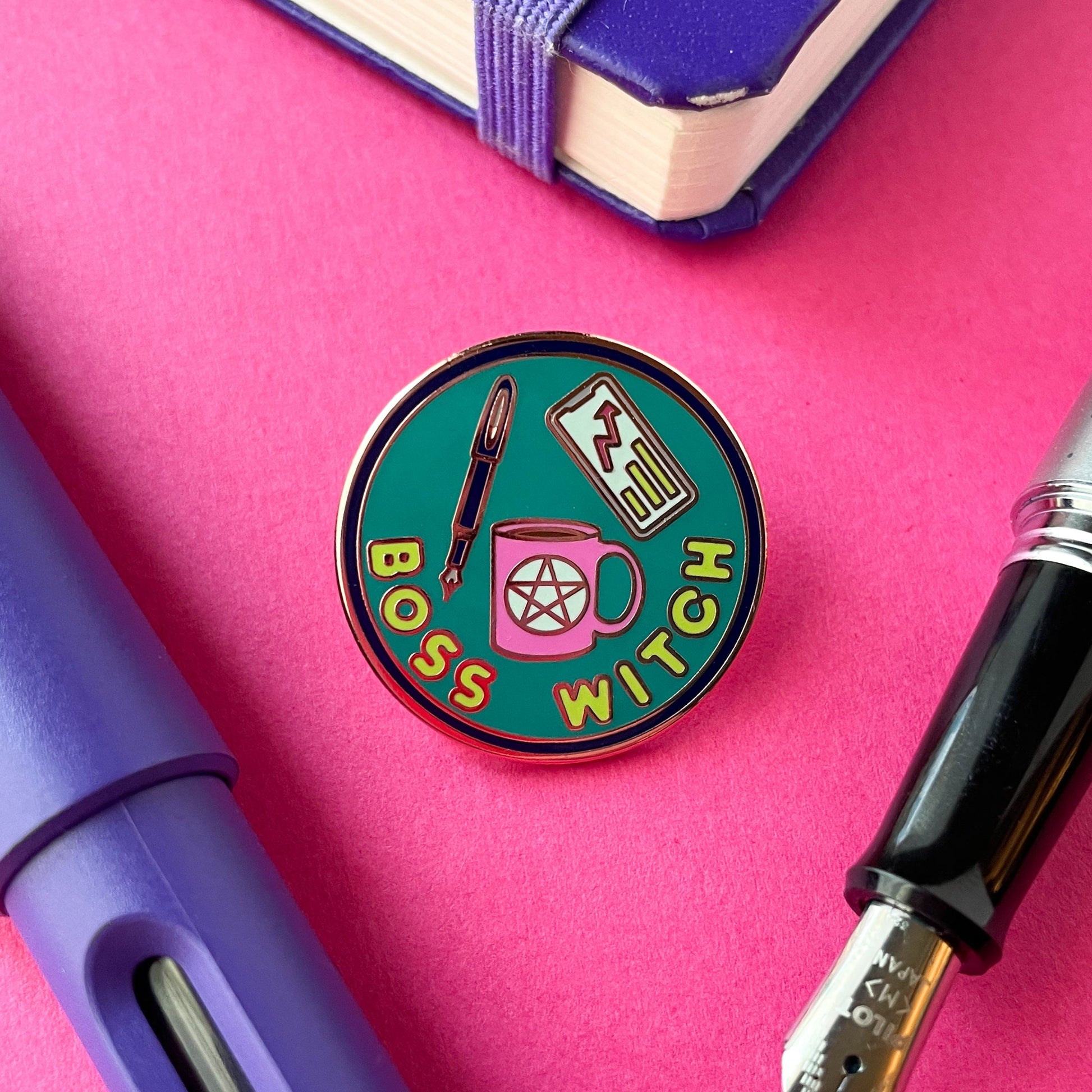 A circular enamel pin on a hot pink background surrounded by fountain pens and a notebook corner. The pin says "Boss Witch" and has a coffee cup with a pentacle, a fountain pen, and a phone with a graph on it depicted on it.