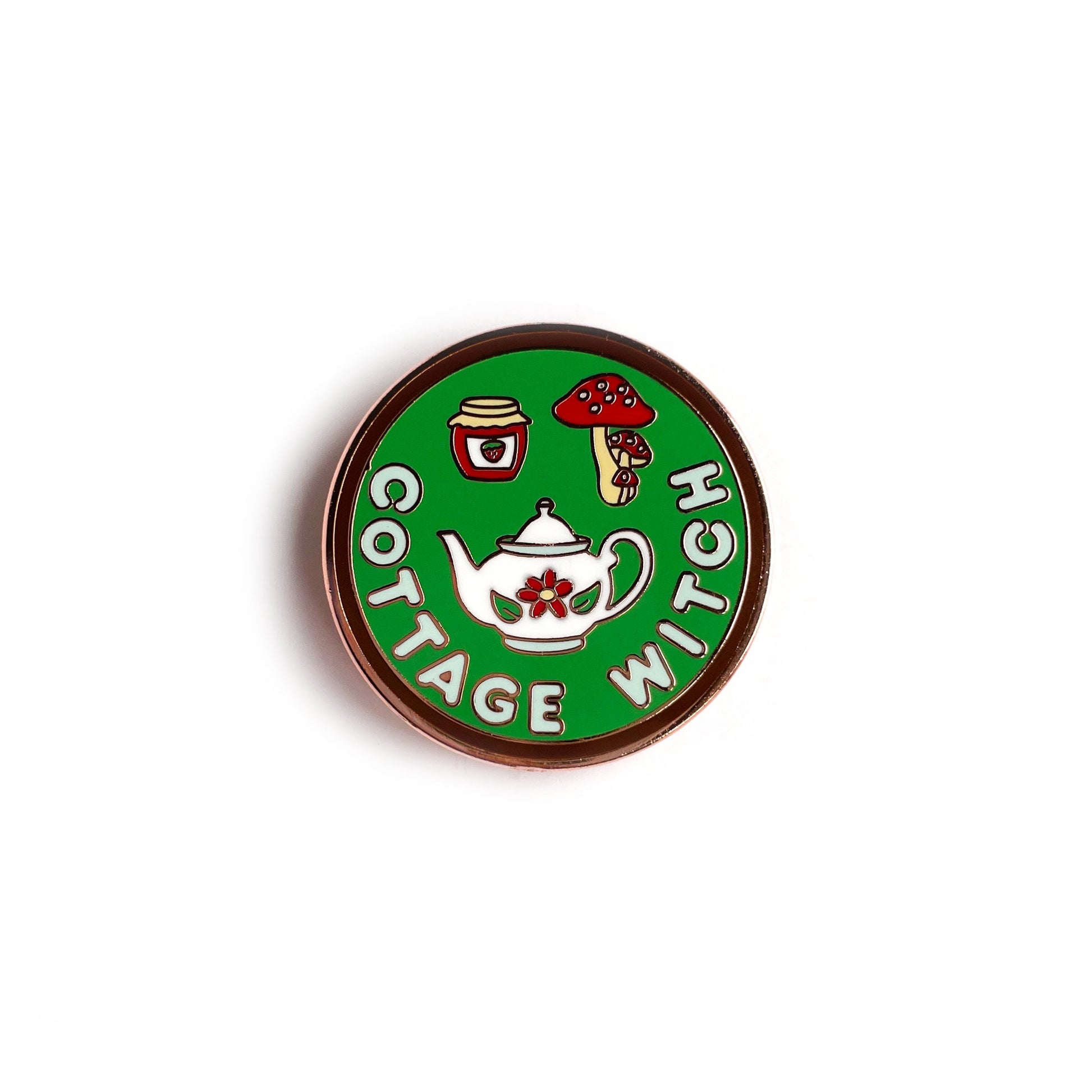 A circular enamel pin. It has a brown border with a sage green background and letters in light blue that read "Cottage Witch". It has a floral teapot, strawberry jam jar, and red mushrooms depicted on it. 