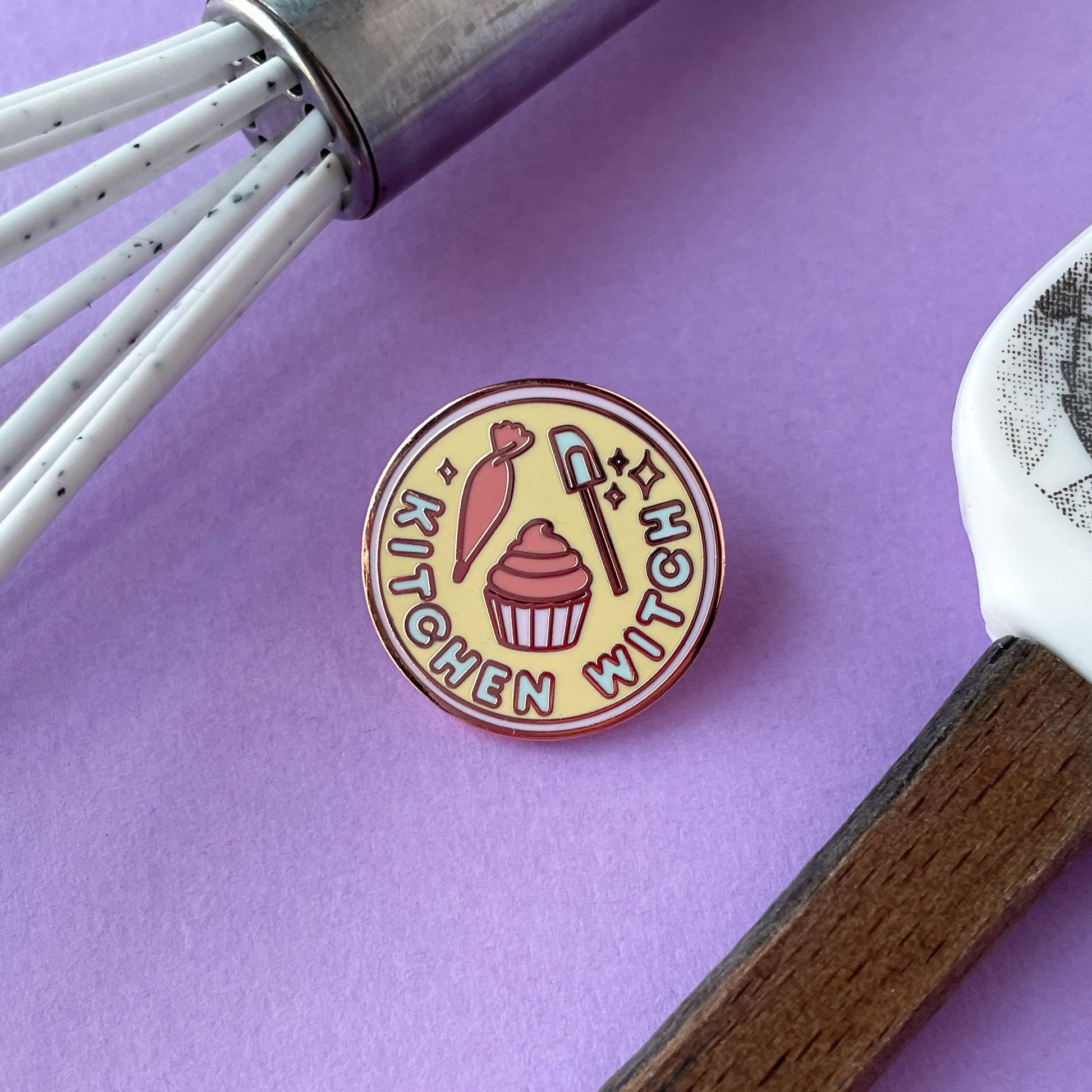 A circular enamel pin on a purple background surrounded by a whisk and spatula handle. The pin has the words "Kitchen Witch" on it. 