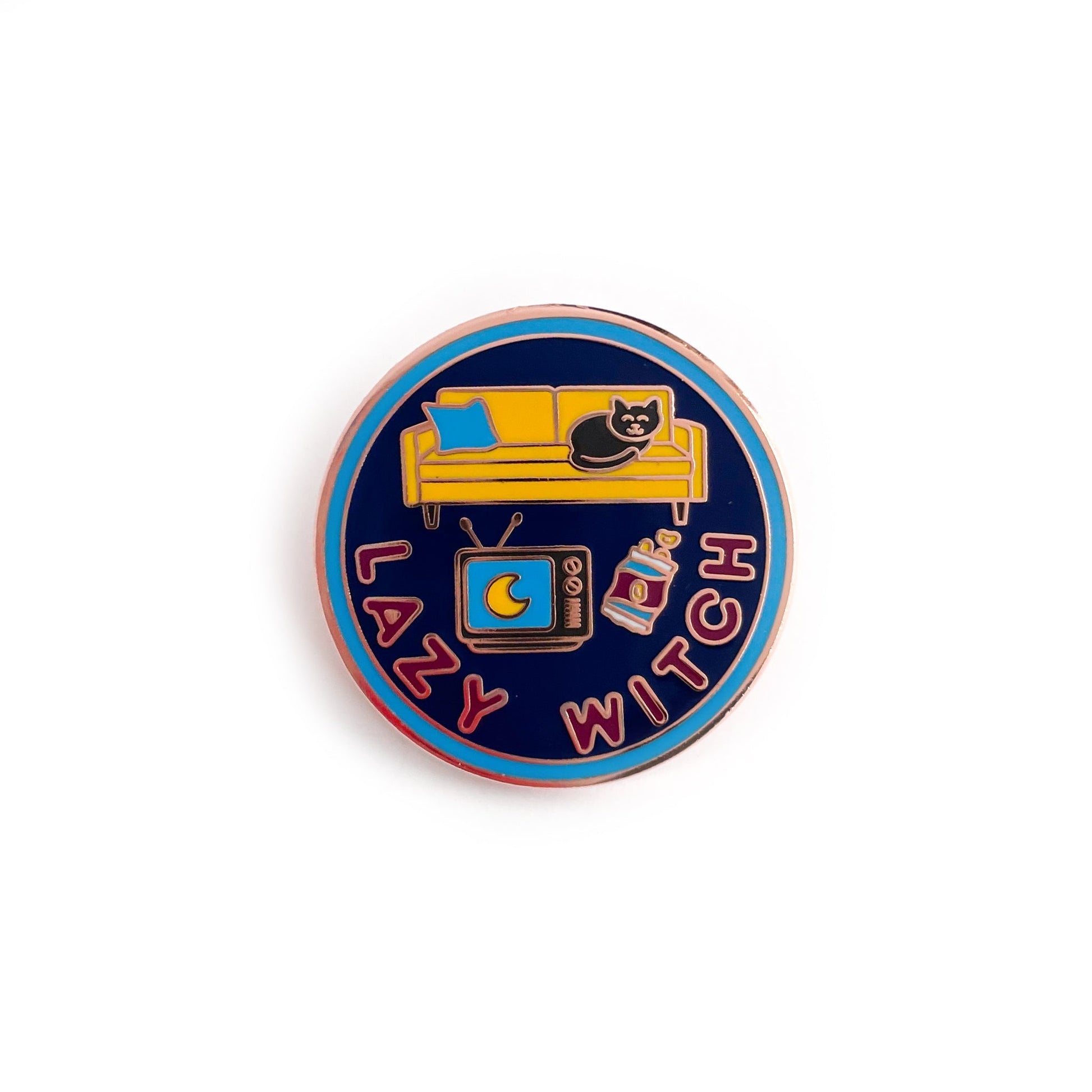 A circular enamel pin with a blue border, navy background, and maroon words that say "Lazy Witch". It has a vintage TV, bag of chips, a yellow couch and a black cat on the couch. 