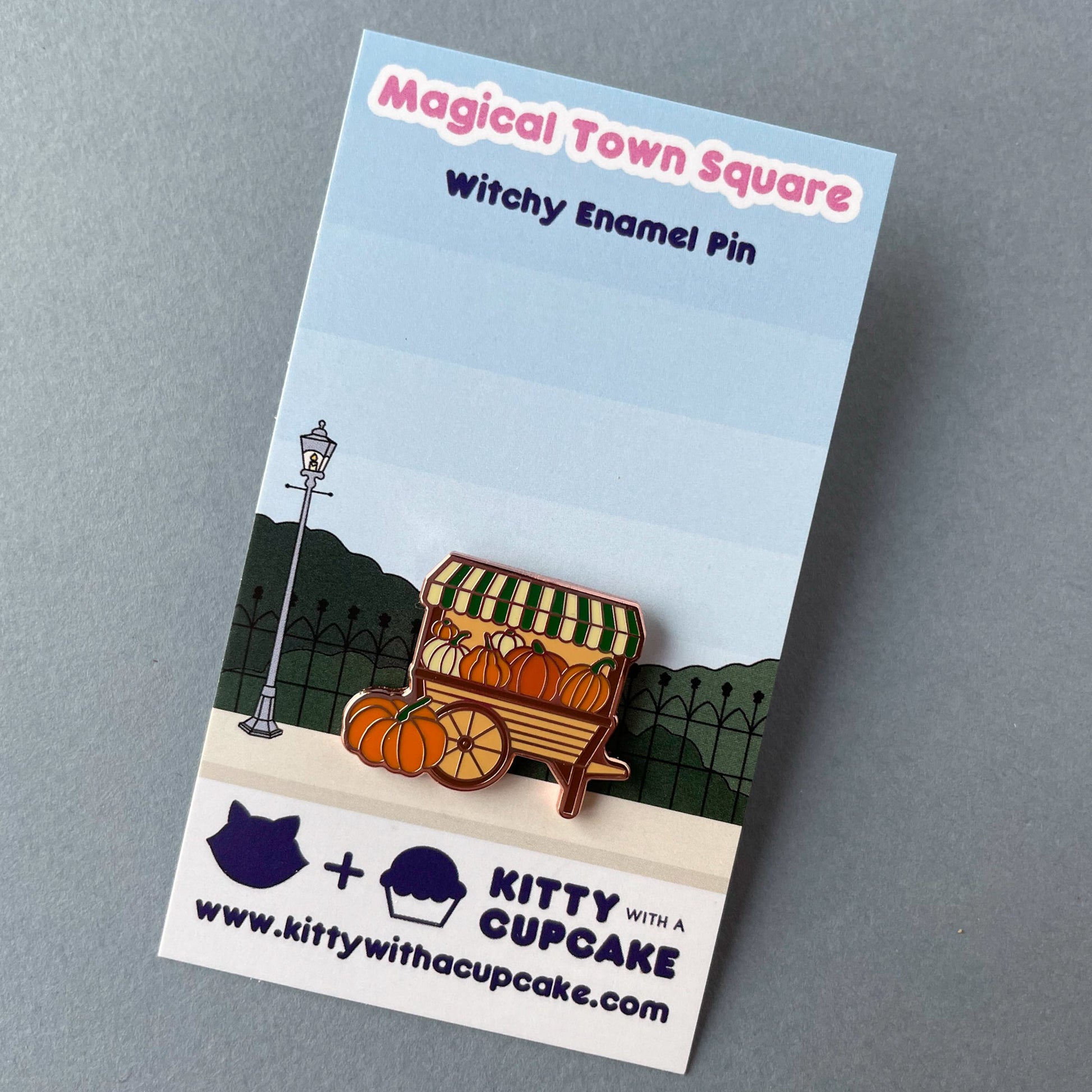A pumpkin cart pin attached to its backing card which reads "Magical Town Square Witchy Enamel Pin" and has a street with a fence and lamp post on it.