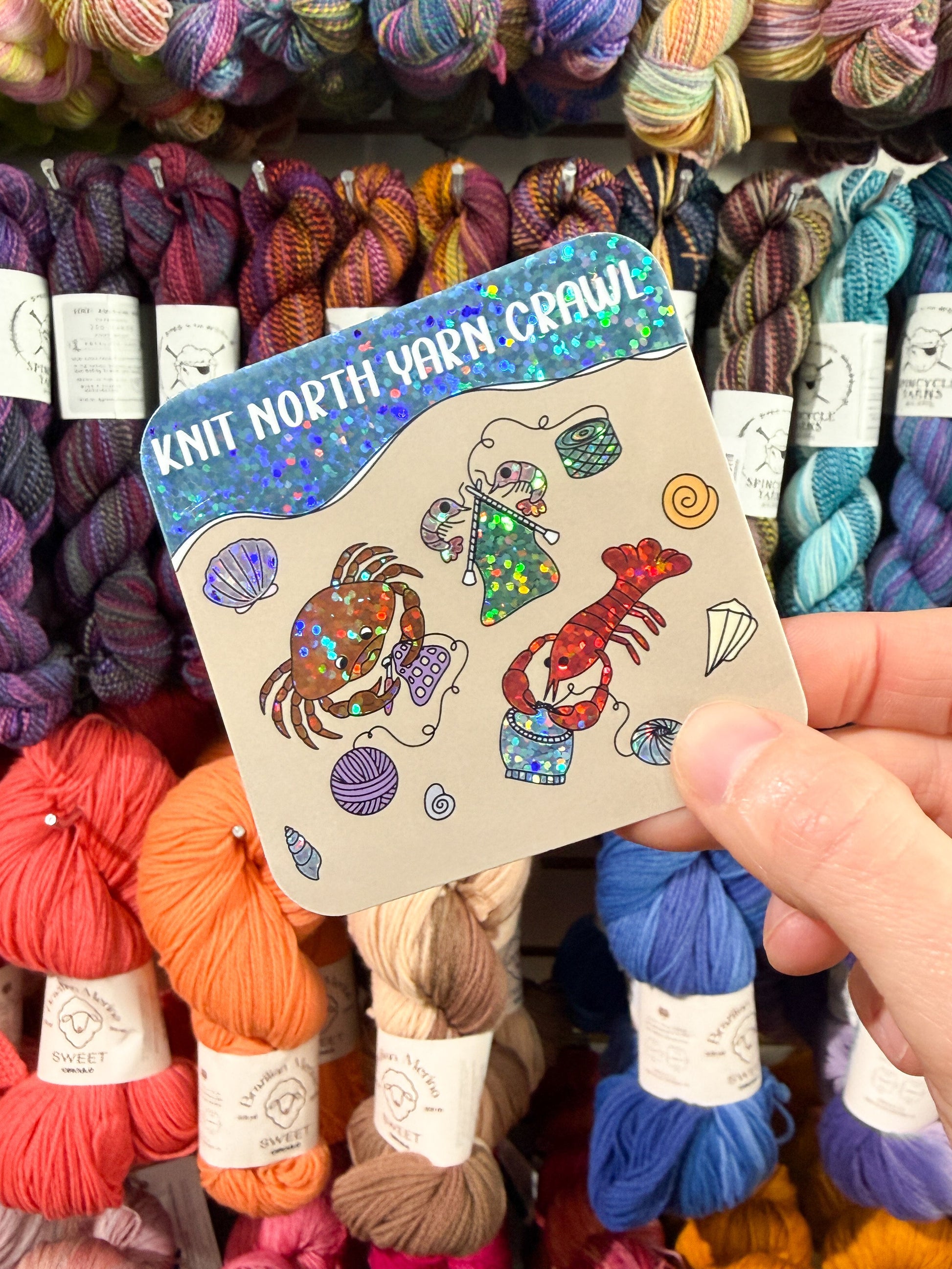 A hand holding a square sticker with rounded edges featuring a crab, lobster, and shrimp knitting and crocheting on a beach with the text 'Knit North Yarn Crawl' on a background of colorful skeins