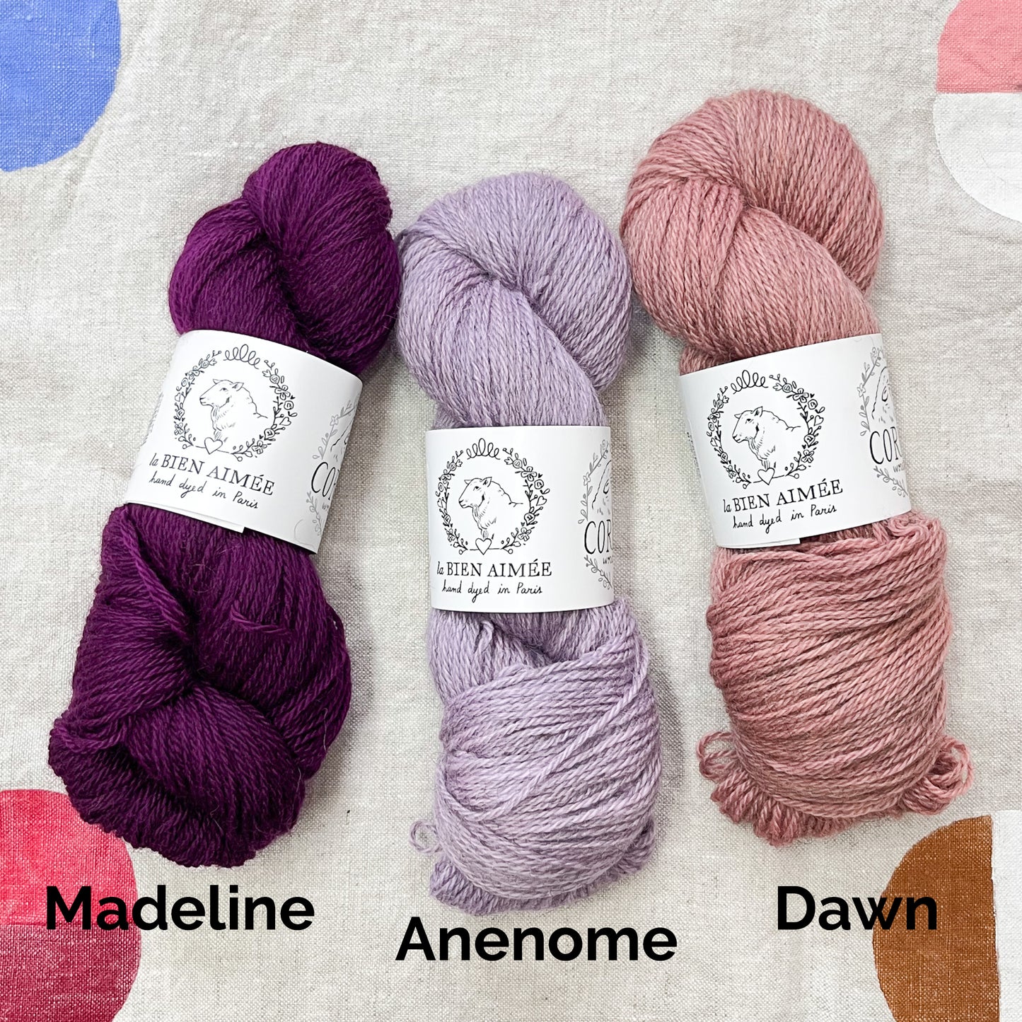 An image of a skein of dark purple yarn on the left, a skein of lilac purple yarn in the middle, and a skein of dusty rose pink yarn on the right, all with white labels featuring an image of a sheep and the title ‘La Bien Aimee’ against a canvas background.