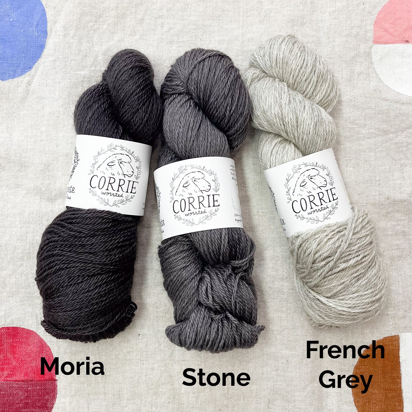 An image of a skein of charcoal gray yarn on the left, a skein of steel gray yarn in the middle, and a skein of heathered pale gray yarn on the right, all with white labels featuring an image of a sheep and the title ‘Corrie Worsted’ against a canvas background.