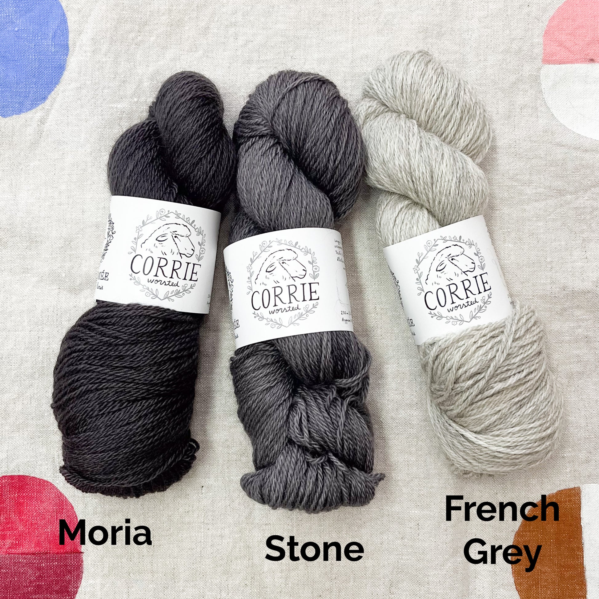 An image of a skein of charcoal gray yarn on the left, a skein of steel gray yarn in the middle, and a skein of heathered pale gray yarn on the right, all with white labels featuring an image of a sheep and the title ‘Corrie Worsted’ against a canvas background.