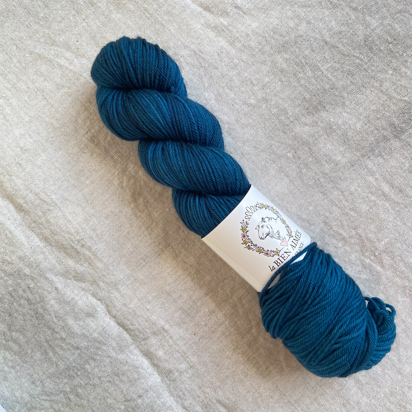 An image of a skein of DK weight yarn in an ocean blue color with a white ‘La Bien Aimee’ label against a canvas background.