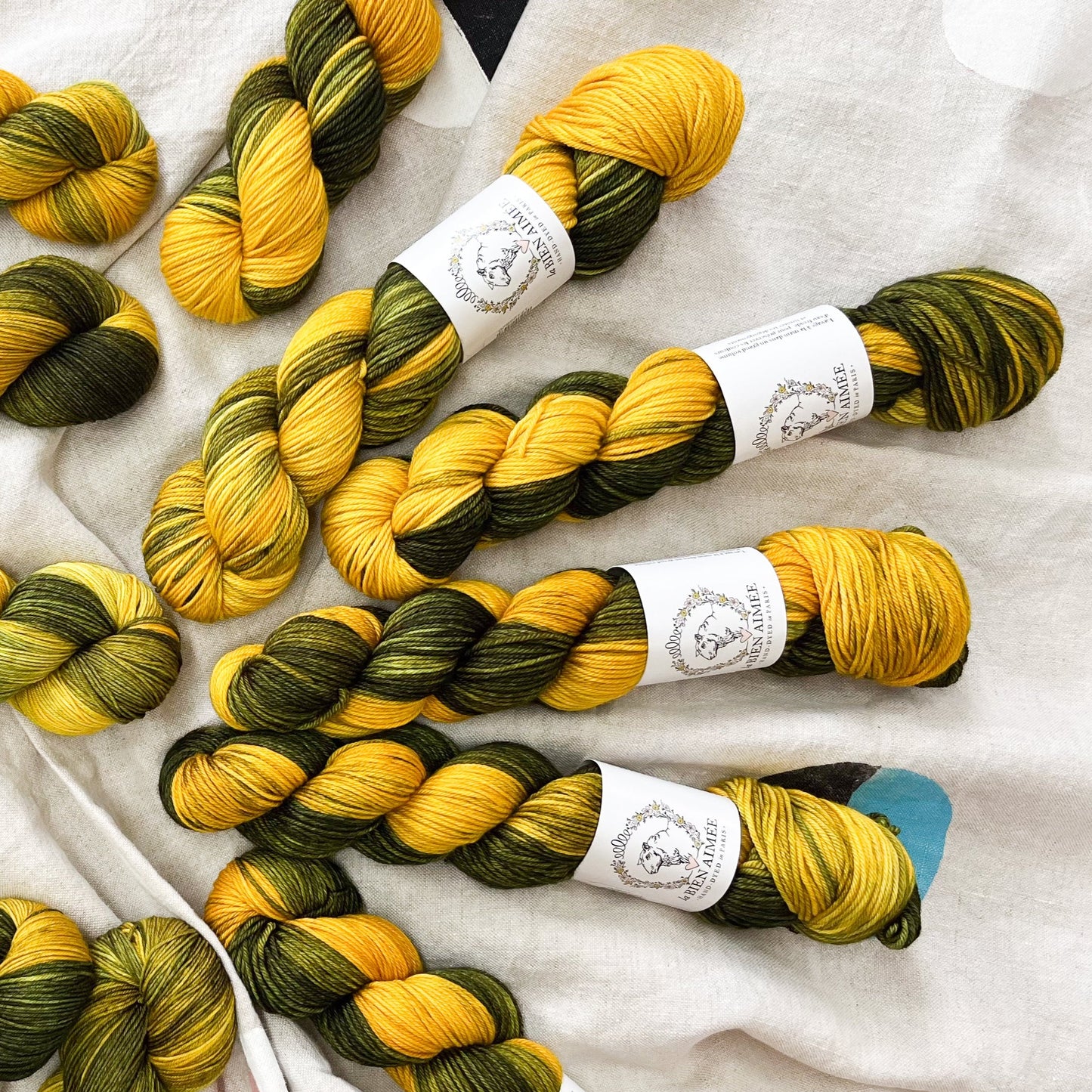 An image of multiple skeins of La Bien Aimee DK weight yarn in shades of dark green and yellow, with white ‘La Bien Aimee’ labels against a canvas background.