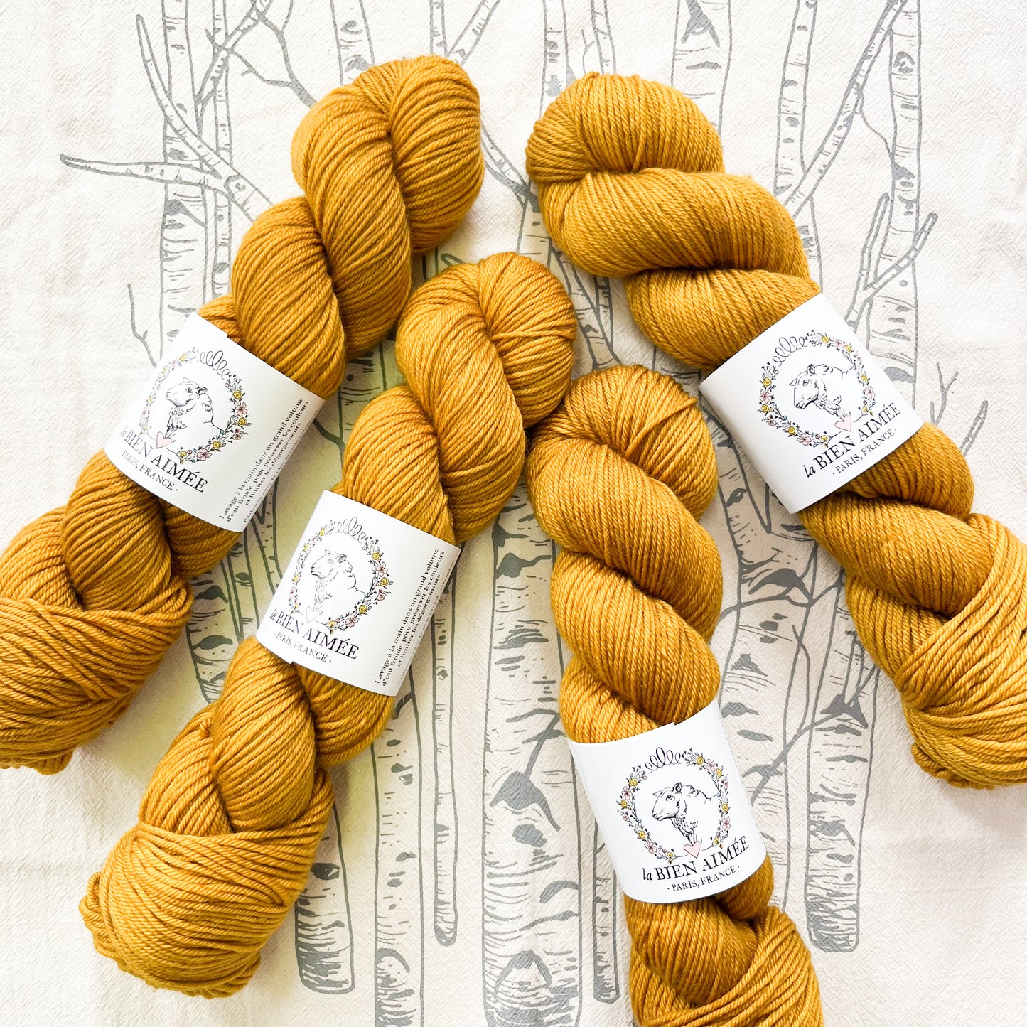 An image of four skeins of DK weight yarn in a mustard yellow color with white  ‘La Bien Aimee’ labels against a white background with tree branch graphics.