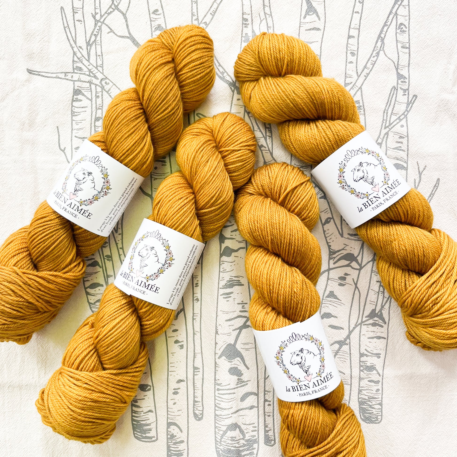 An image of four skeins of DK weight yarn in a mustard yellow color with white  ‘La Bien Aimee’ labels against a white background with tree branch graphics.
