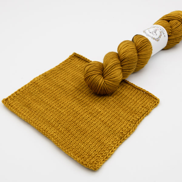 An image of a skein of DK weight yarn in a mustard yellow color with a white ‘La Bien Aimee’ label, laying on a yellow yarn swatch against a white background.