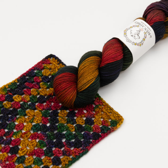 An image of a skein of DK weight yarn in dark shades of blue, green, yellow, orange, and purple with a white ‘La Bien Aimee’ label lying on a crocheted yarn swatch, against a white background.