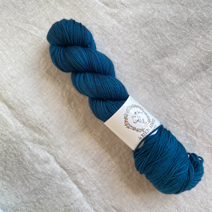 An image of a skein of sock yarn in an ocean blue color, with a white ‘La Bien Aimee’ label against a canvas background.