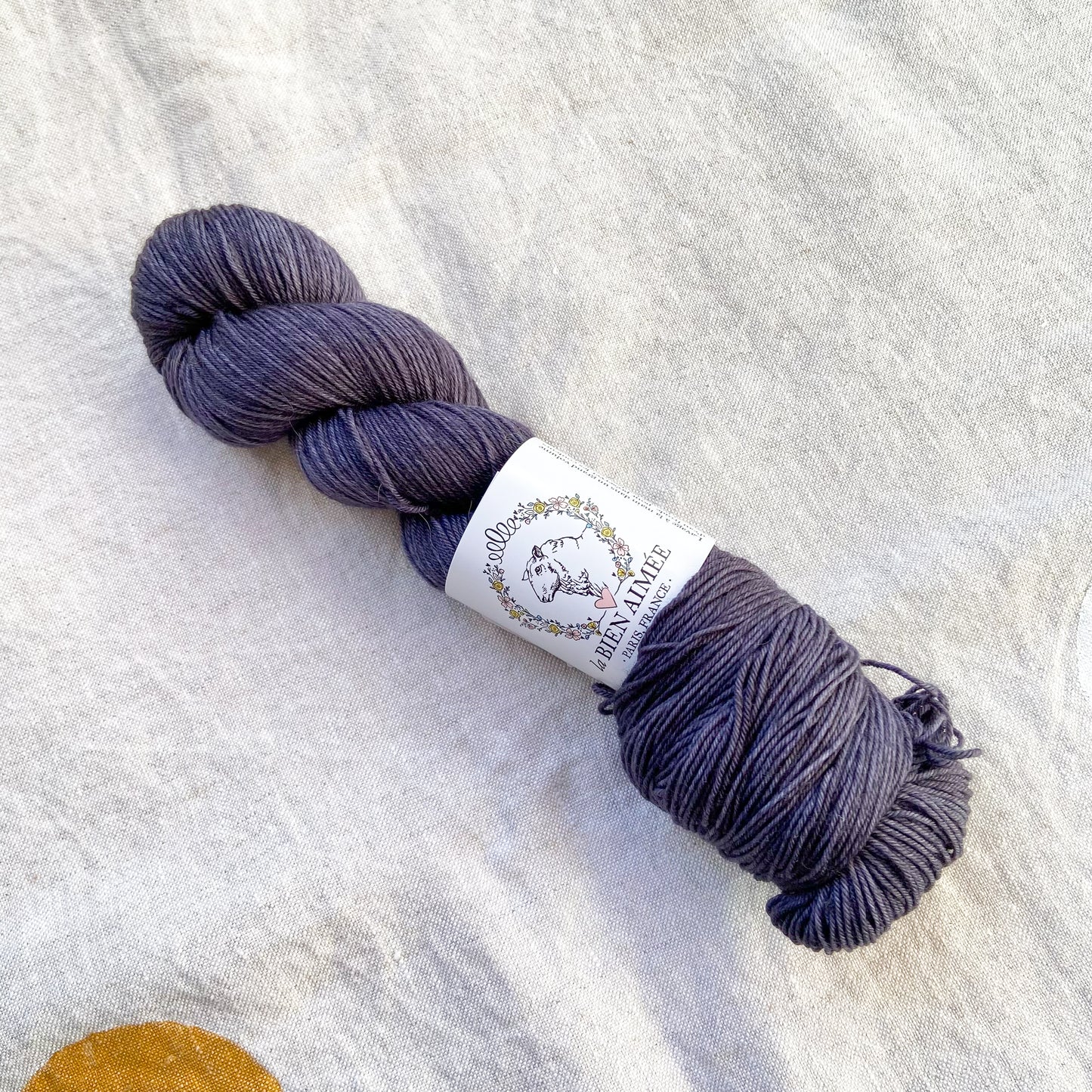 An image of a skein of sock yarn in a charcoal gray color with a white ‘La Bien Aimee’ label against a canvas background.