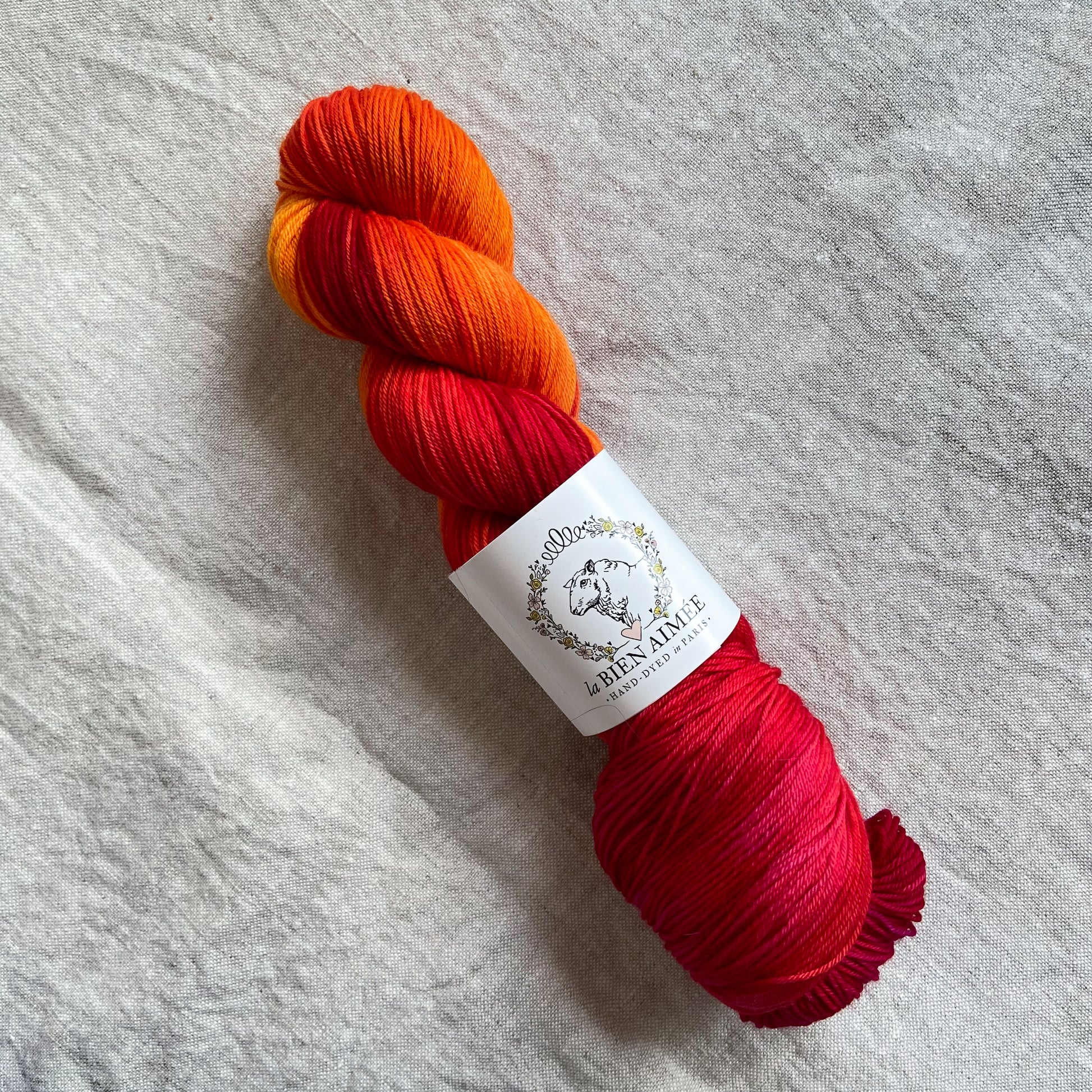An image of a skein of sock yarn in vibrant shades of red and orange,  with a white ‘La Bien Aimee’ label against a canvas background.