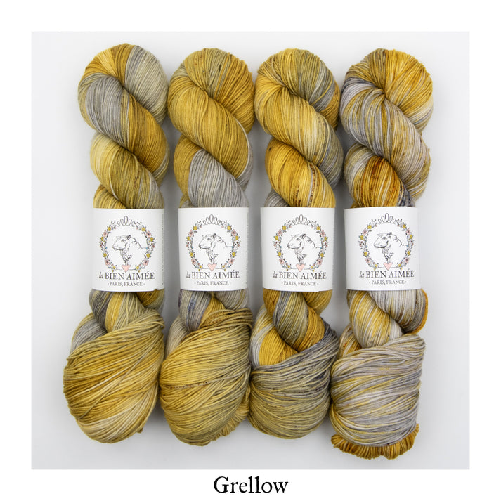 An image of four skeins of sock yarn in shades of yellow and gray with white ‘La Bien Aimee’ labels against a white background.