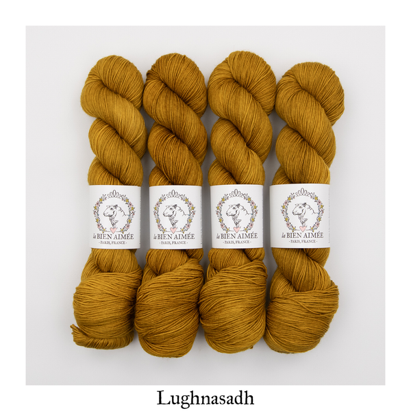 An image of four skeins of La Bien Aimee sock yarn in a mustard yellow color with a white ‘La Bien Aimee’ label against a white background.